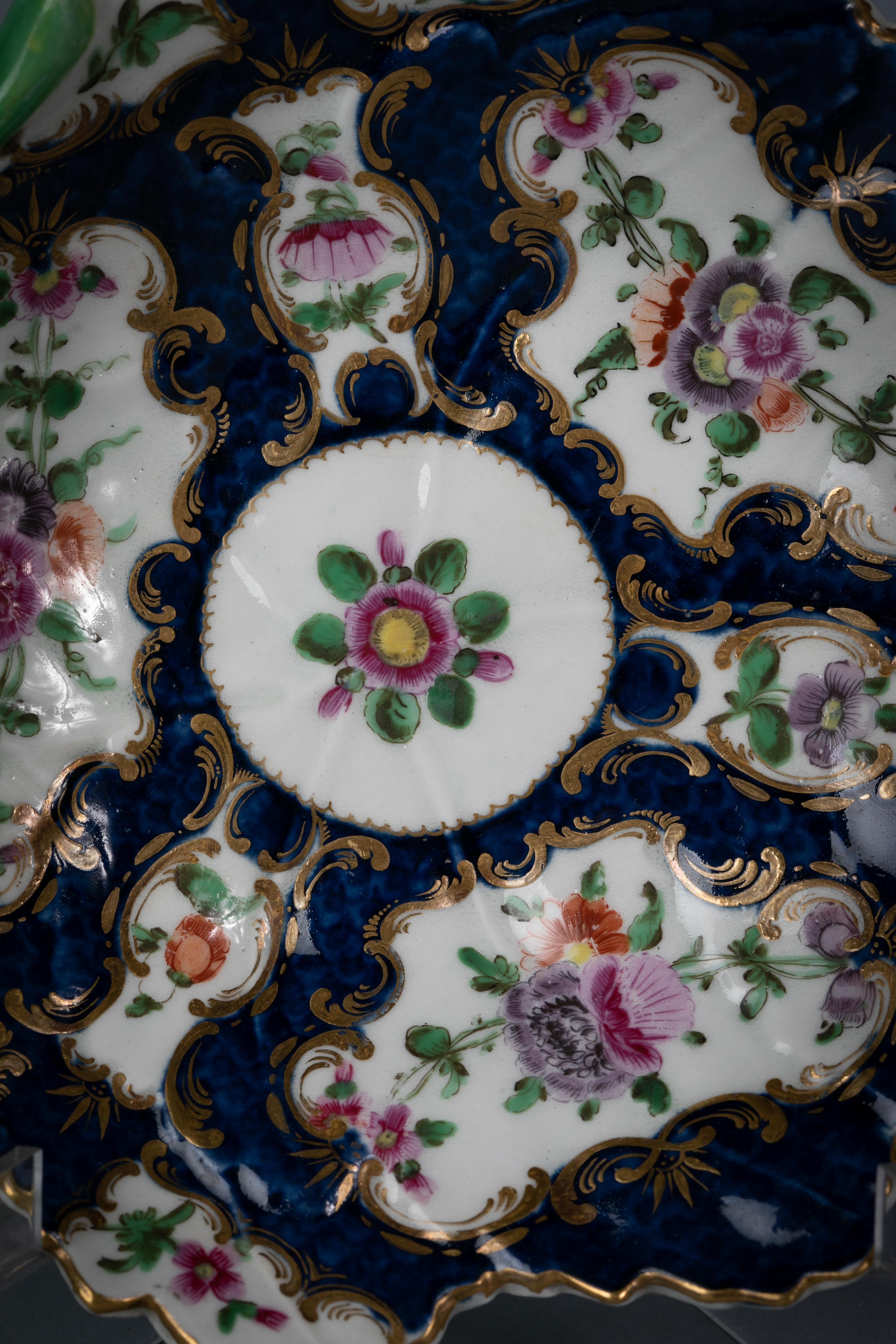 With a twig handle, painted with flowers in reserve panels, edged with gilt, upon a blue scale ground. The shape is derived from a Meissen original. The mark is a fretted square.