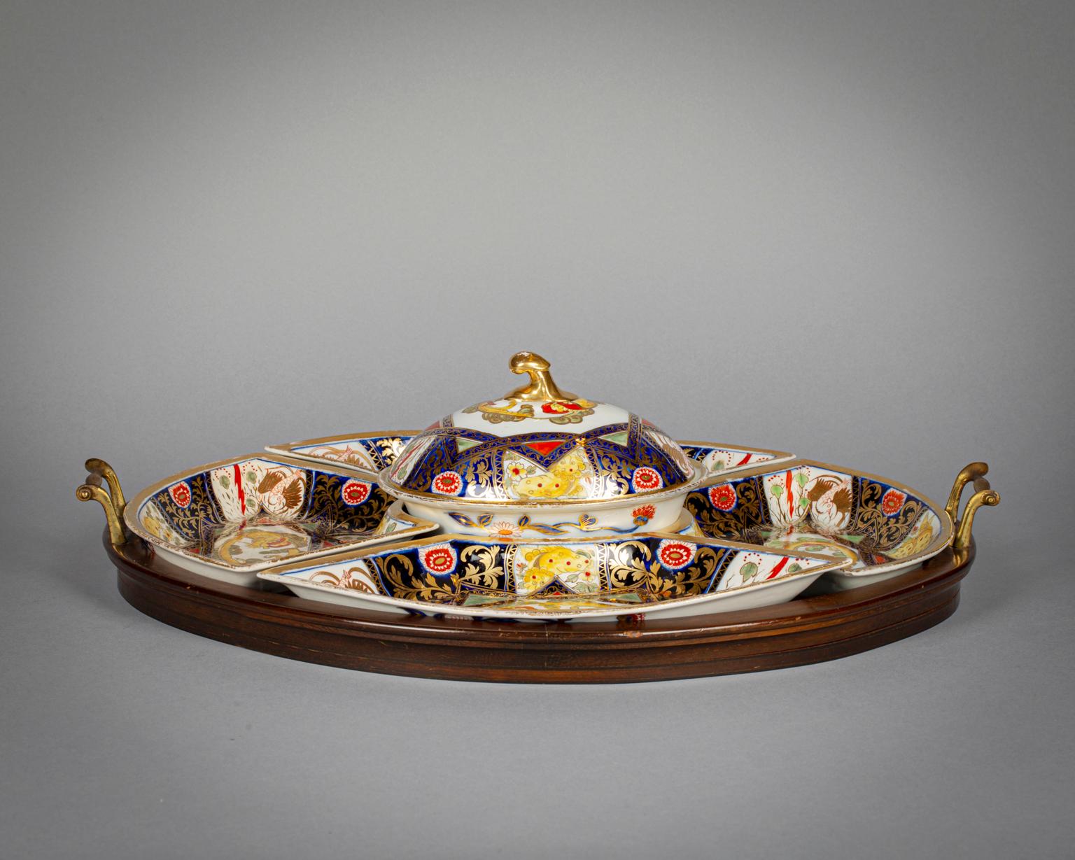 Painted in a variation of the dragon-in-compartment pattern in Imari colors. Consisting of four dishes surrounding a covered tureen resting on a wooden and brass-handled tray. Chamberlain Worcester or Coalport.