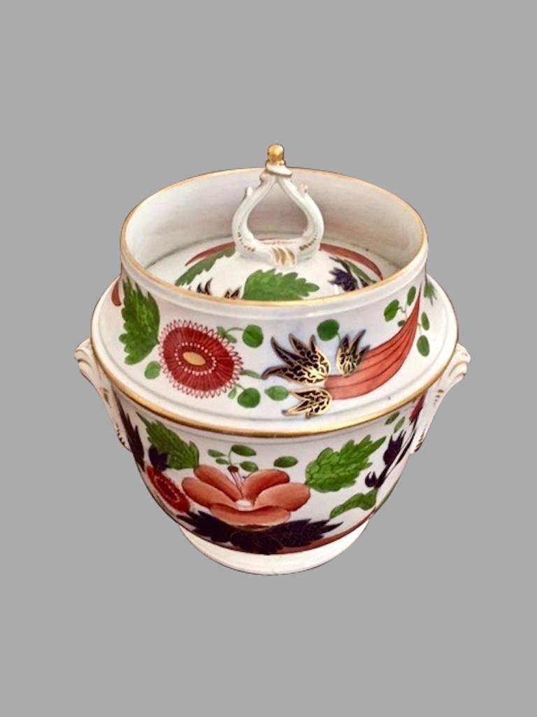 A boldly decorated English covered fruit or ice cooler in the Chinese Imari pattern, decorated in underglaze blue, iron red and gold with chrysanthemums and peonies, the top with a gilt open flame finial, the base with gilded side handles. Retains a