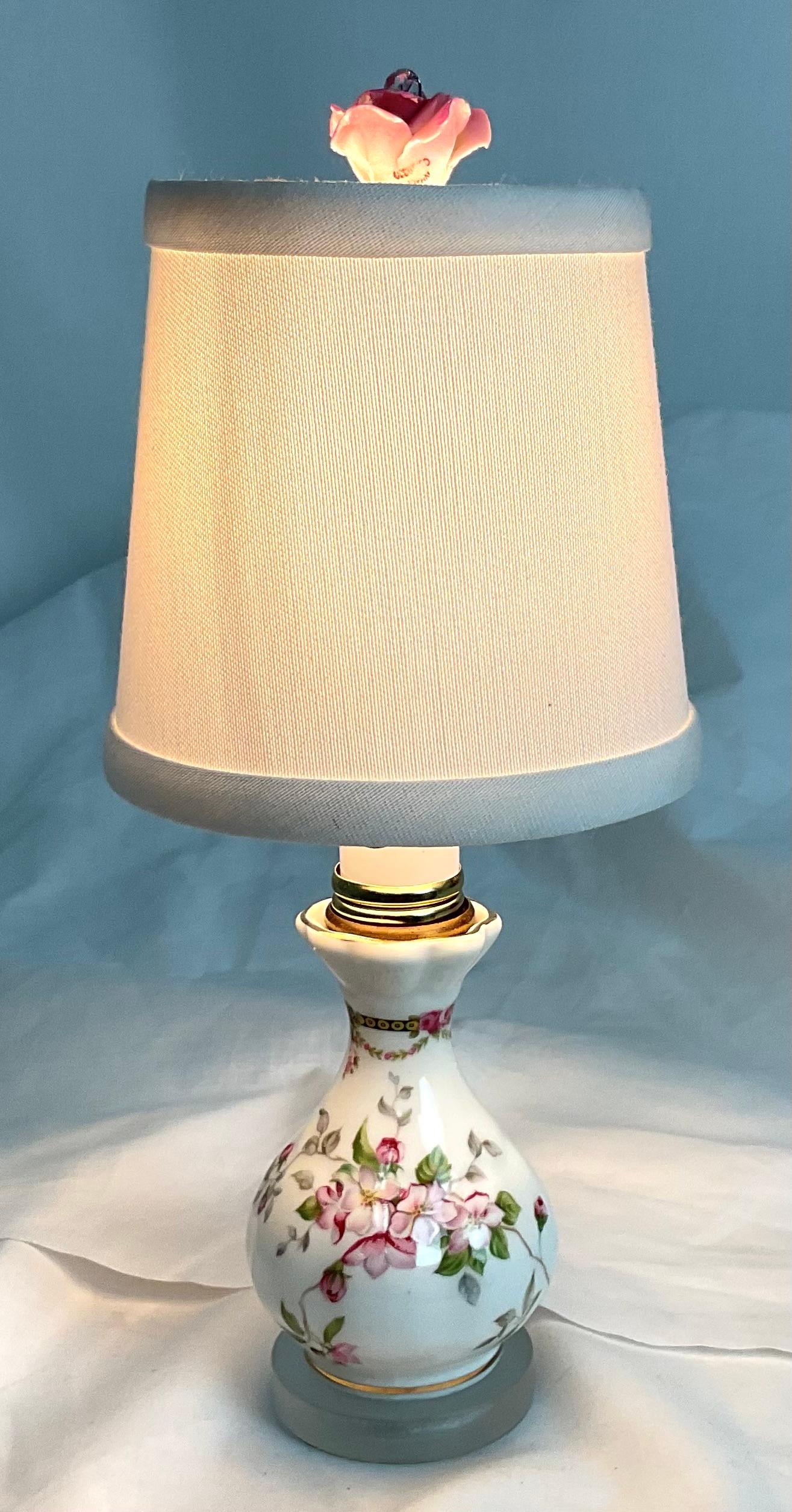 A charming accent piece that embodies timeless elegance, this vintage porcelain table lamp features a delicate hand-painted floral motif in soft pinks and greens on a glossy white base. The graceful silhouette is complemented by gold trim detailing,