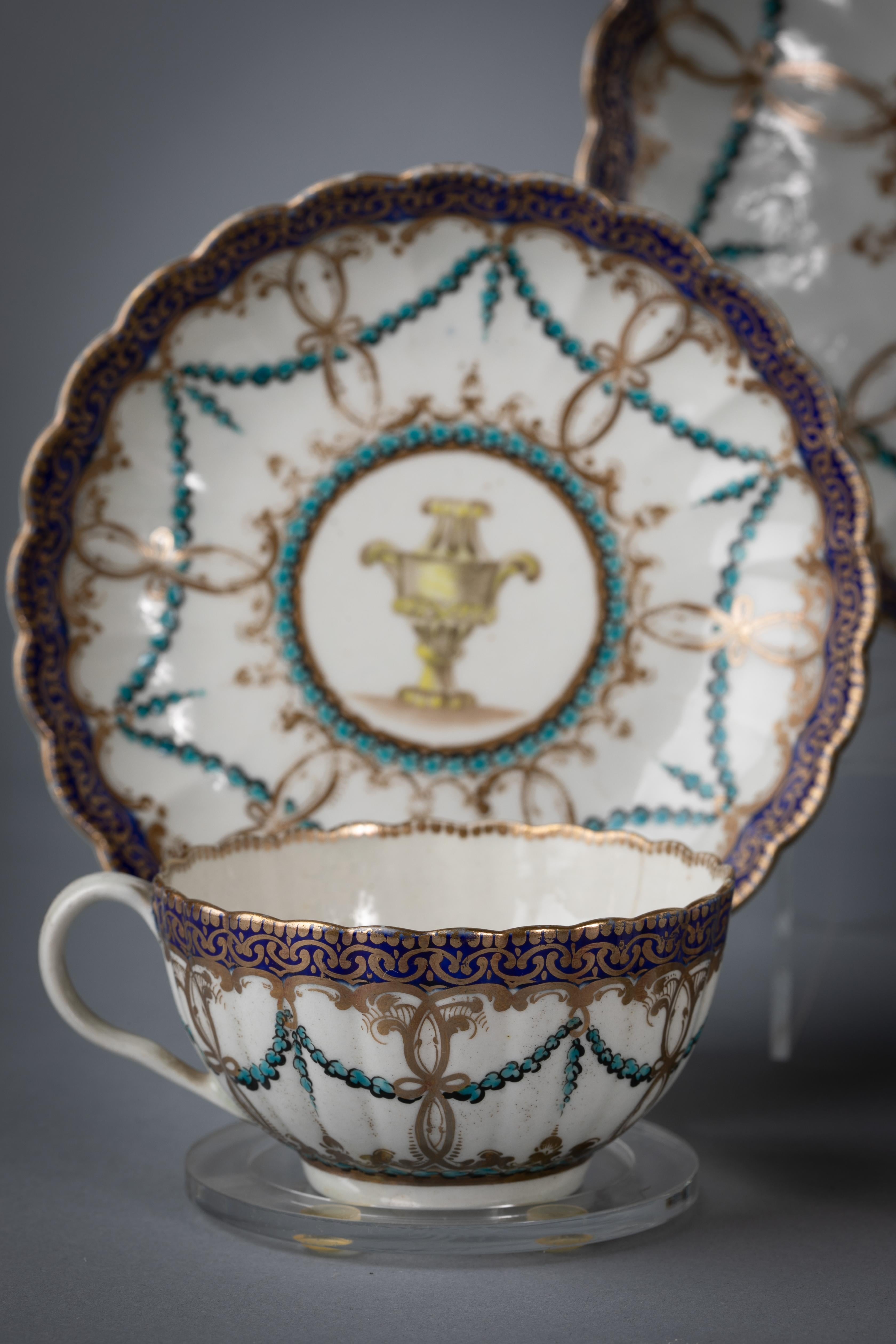 Delicately enameled with turquoise garlands draped through hanging gilt rings beneath a royal blue and gilt border. Each piece with a neoclassical vase painted in grisaille and light yellow. Comprising a bowl, creamer, covered sugar, tea stand, pair
