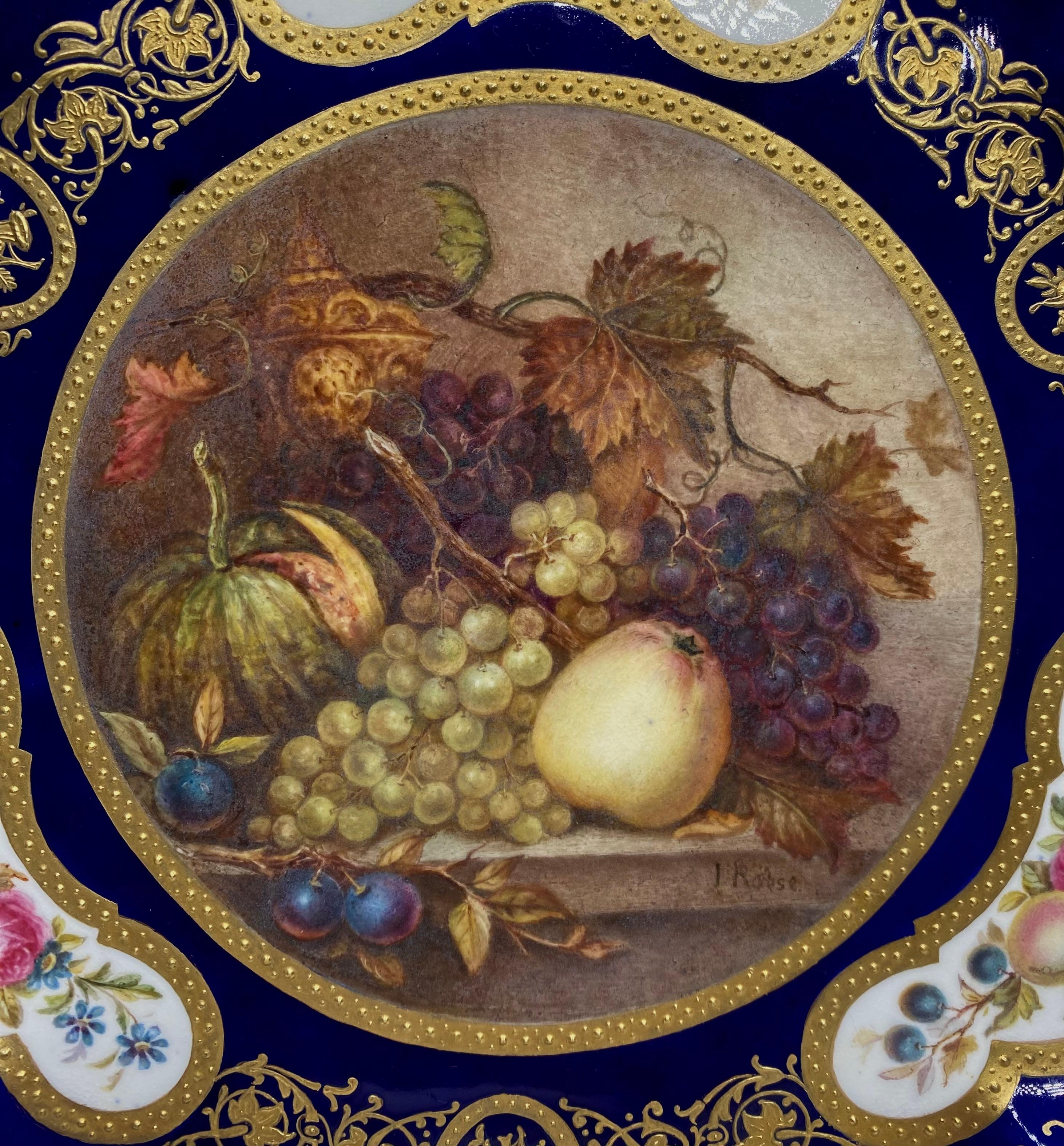 A fine English porcelain plate, painted by James Rouse jnr. c. 1880. Beautifully painted by this exceptional artist, with a circular panel containing a still life of fruit, and a gilt bronze vase, resting upon a marble ledge.
The border with three