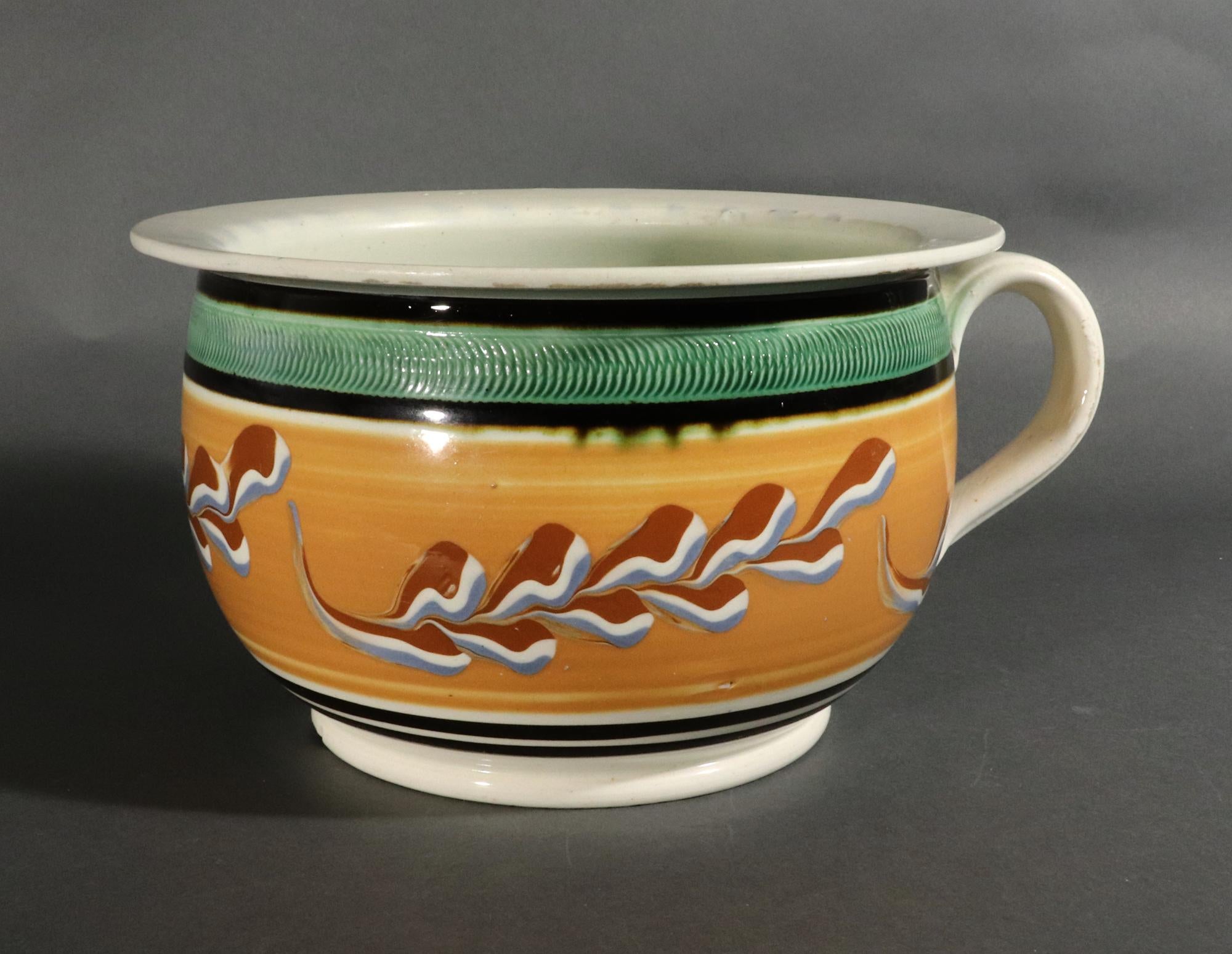 English Pottery Mocha Leaf Pattern With A Light Ochre Band Chamber Pot
Circa 1820

Chamber pots are a rare form in period mocha.  This striking example is of circular form with the body slip-decorated with a ochre ground with tri-color leaves around