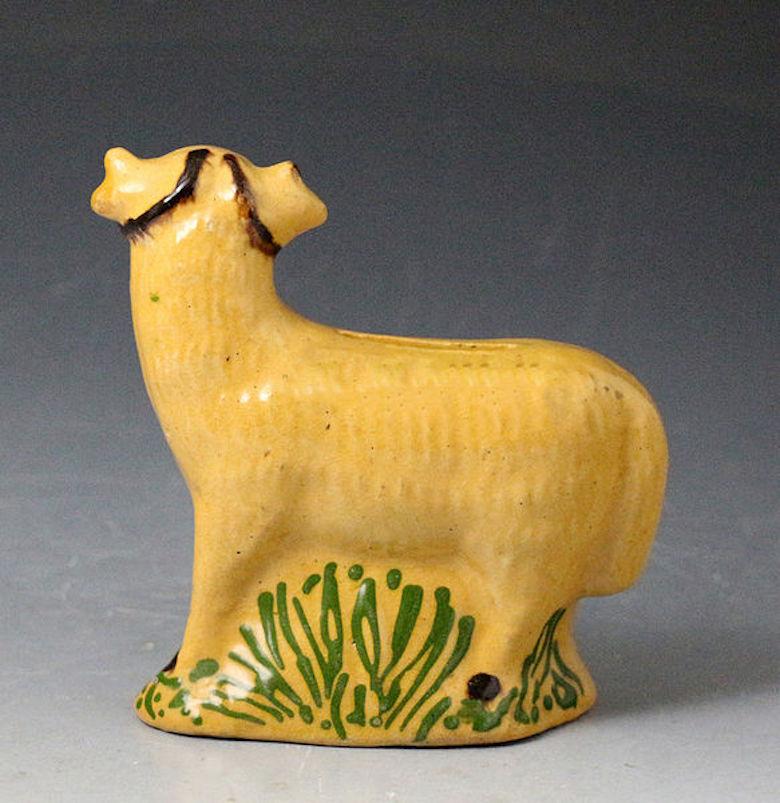 A naive buff-colored clay pottery figure of a ram in the form of a money box bank. The piece is modeled standing on a grassy base decorated in an abstract manner. Probably North Country England, 1825. 

Medium: earthenware pottery
