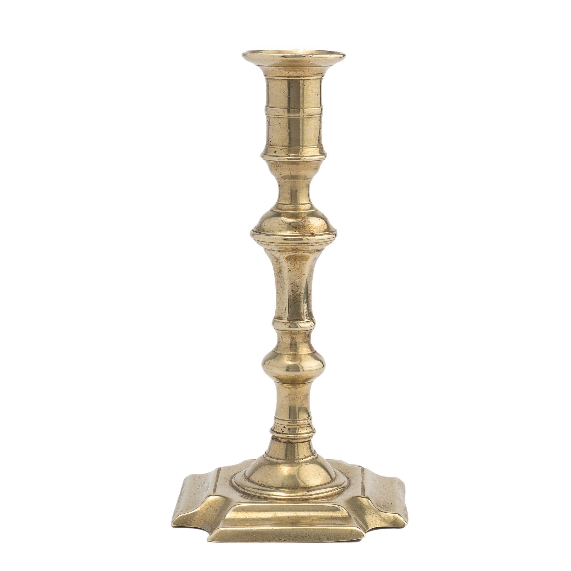 Queen Anne brass candlestick with a hollow core cast baluster. The bluster is cast with a candle cup with medial ring along with a cupped circular bobeshe. The baluster is peened to a circular dome on a raised square base with cove cut