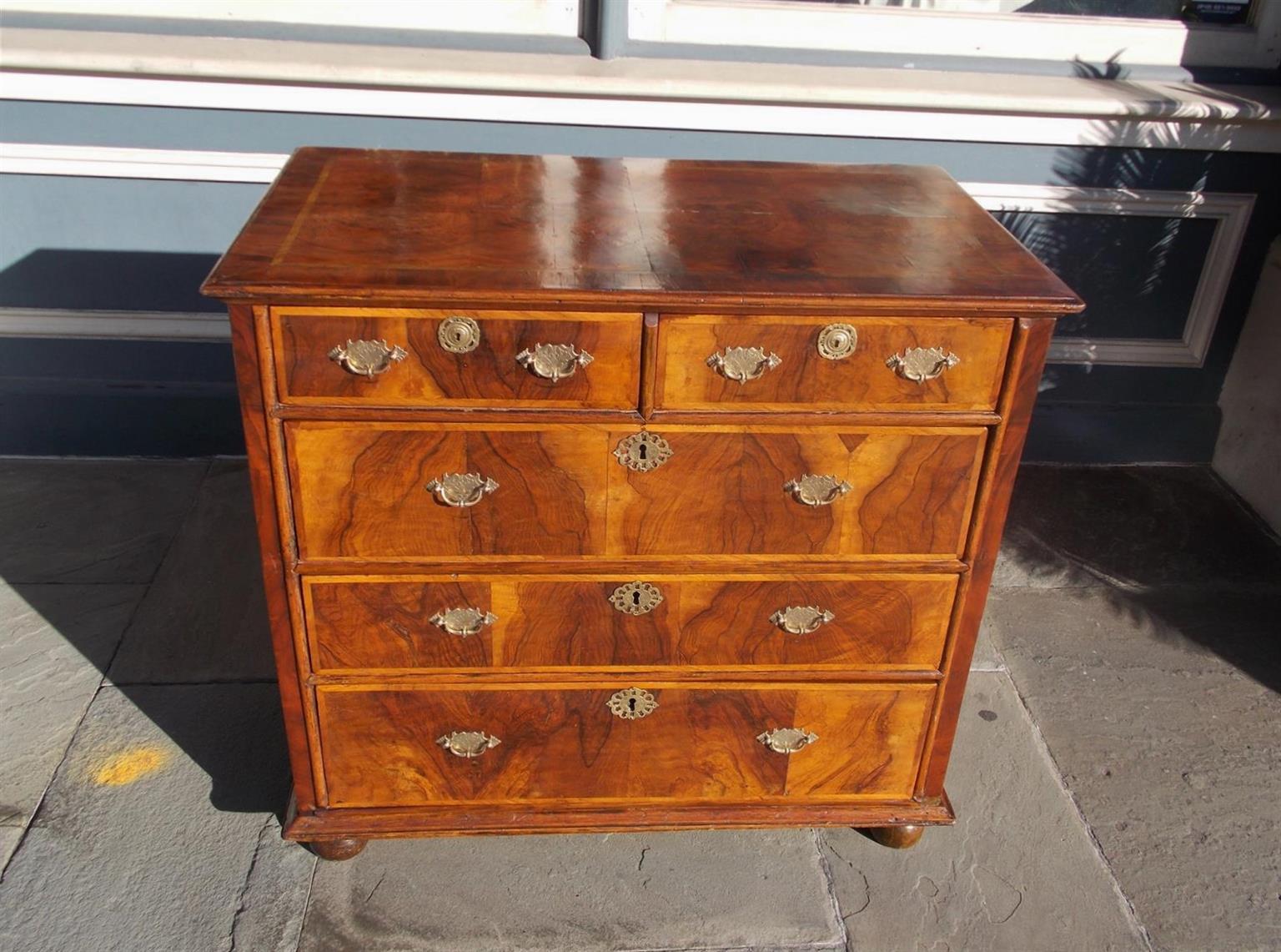 English Queen Anne book matched burl walnut five-drawer chest with a carved molded edge, original brasses and escutcheons, feather banded satinwood inlays, and resting on bun feet, Mid-18th century.