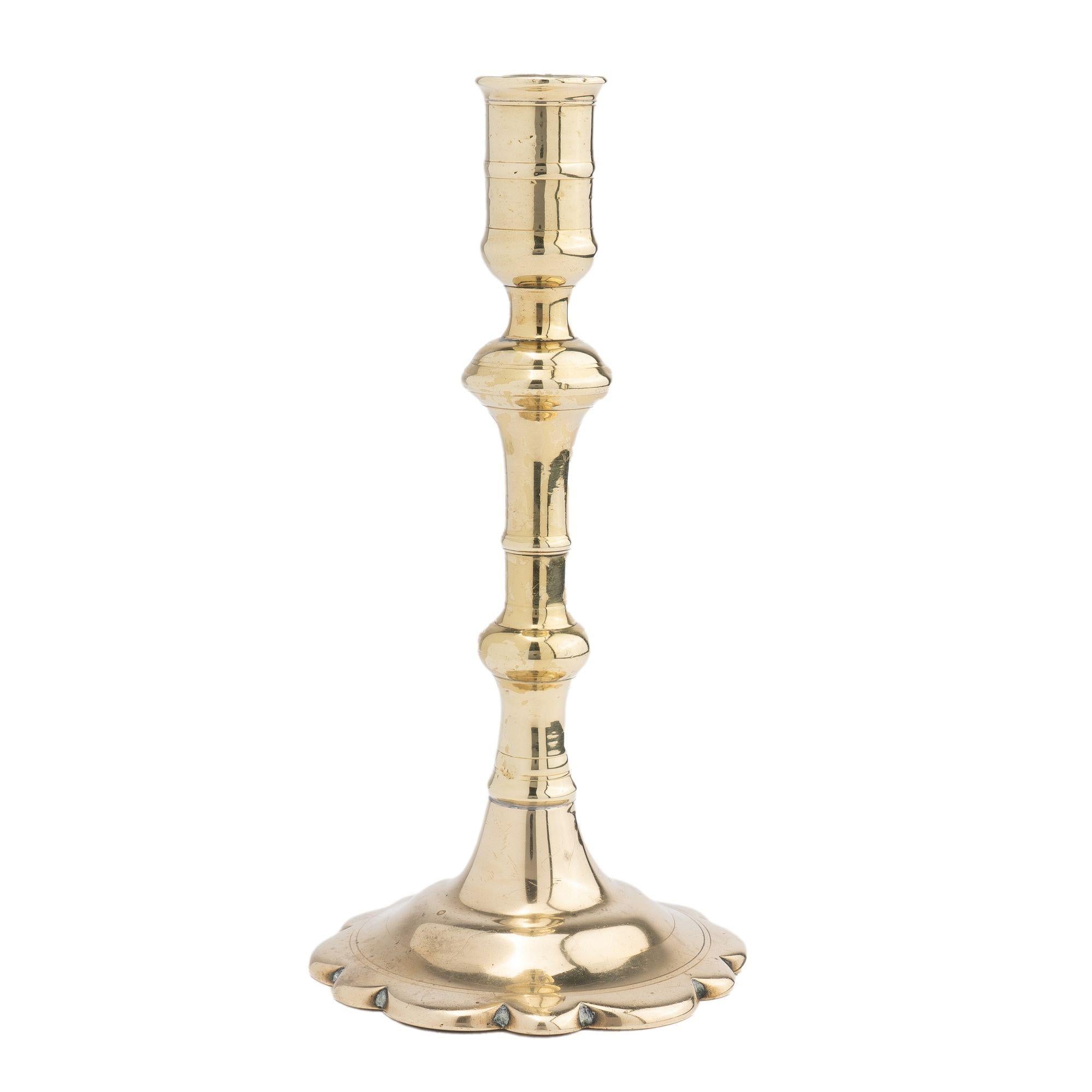Seam cast brass Queen Anne candlestick with urn form candle cup supported by a trumpet form shaft over a suppressed knob peened to a conical to domed base with scolloped edge. The candle shaft retains its original push up candle
