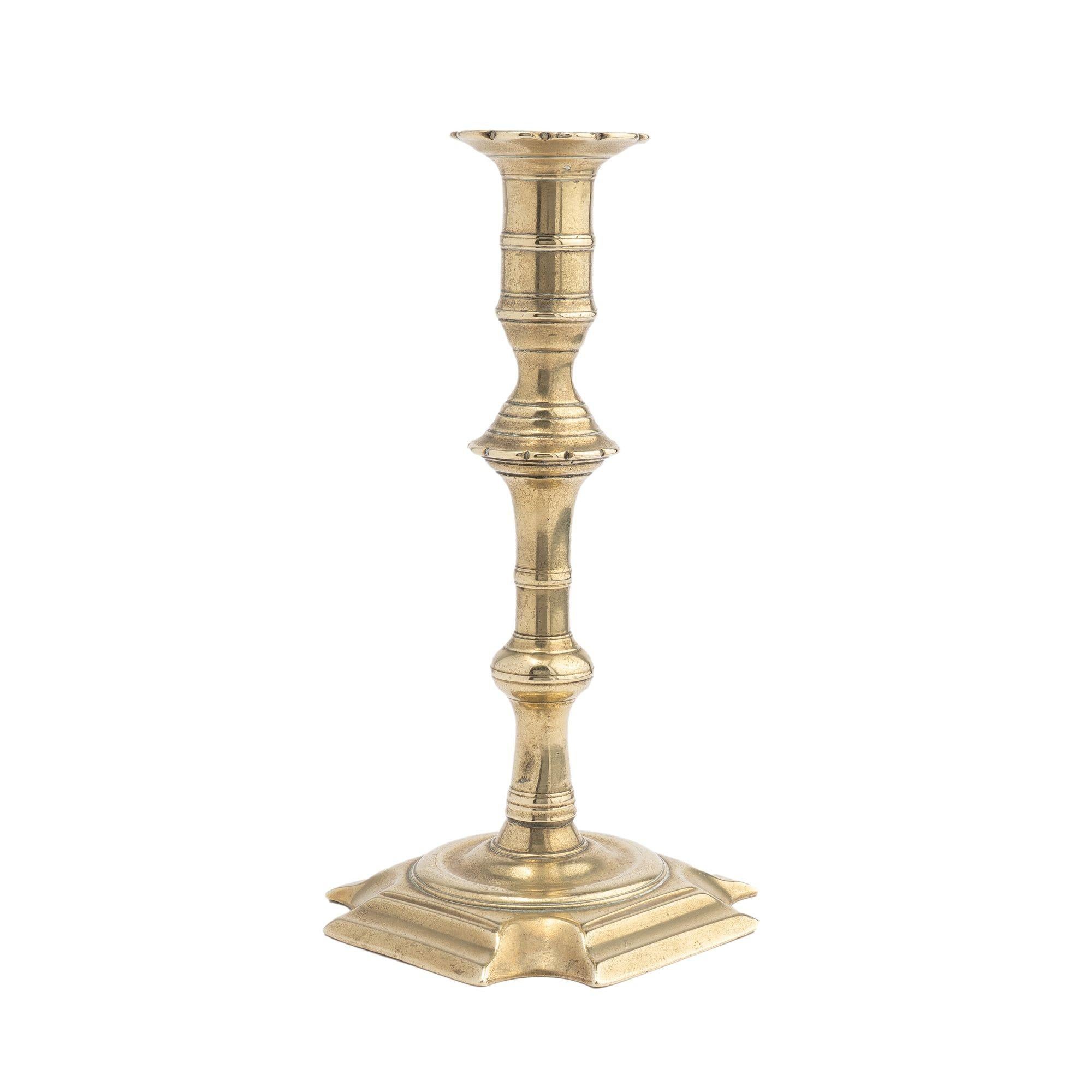 Core cast brass Queen Anne baluster candlestick. The candlestick is cast with a scolloped edge bobeshe and tall candle cup with bead moldings above a scolloped edge, knob beaded lower shaft, and lower knob. The base is a shallow square around a