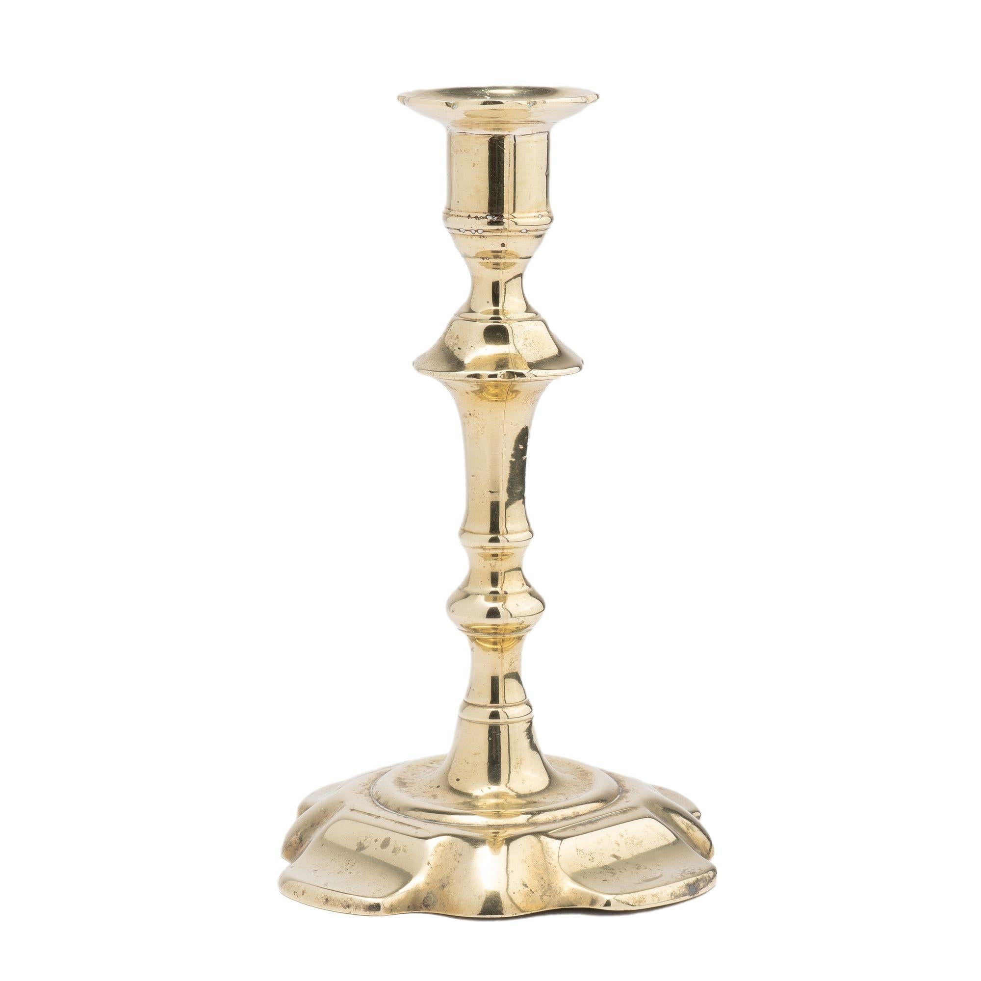 Seam cast brass Queen Anne candlestick. The candlestick features an urn form candle cup with scalloped bobeche on a trumpet turned shaft. The suppressed knob is peened to a cone centered on a shallow dished circle resting on an extended petal