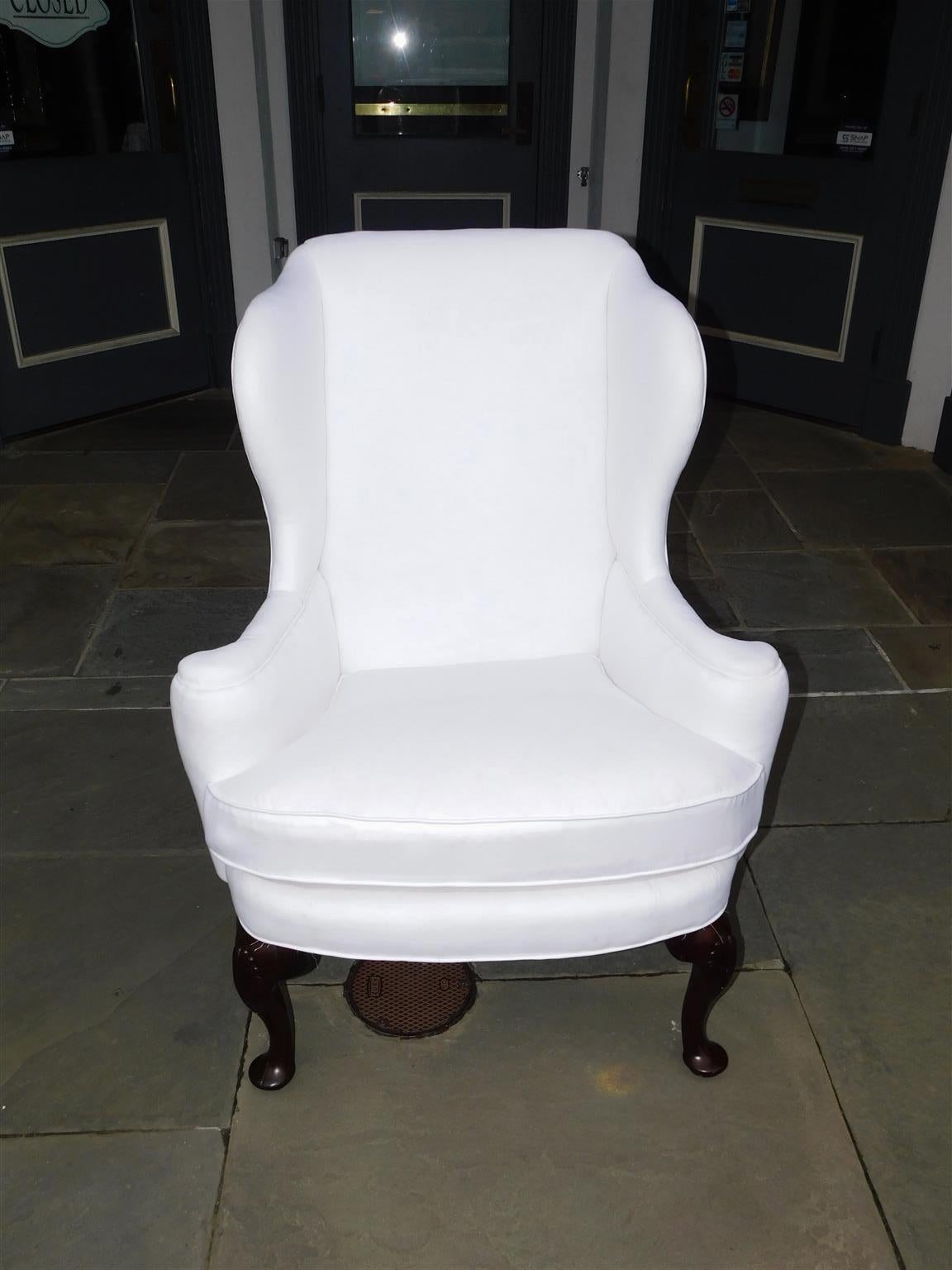 English Queen Anne Mahogany upholstered wingback chair with flanking flared wings, flanking scrolled arms, carved shell knees, and resting on the original pad feet with rear squared tapered legs. Mid-18th century. wingback chair is upholstered in
