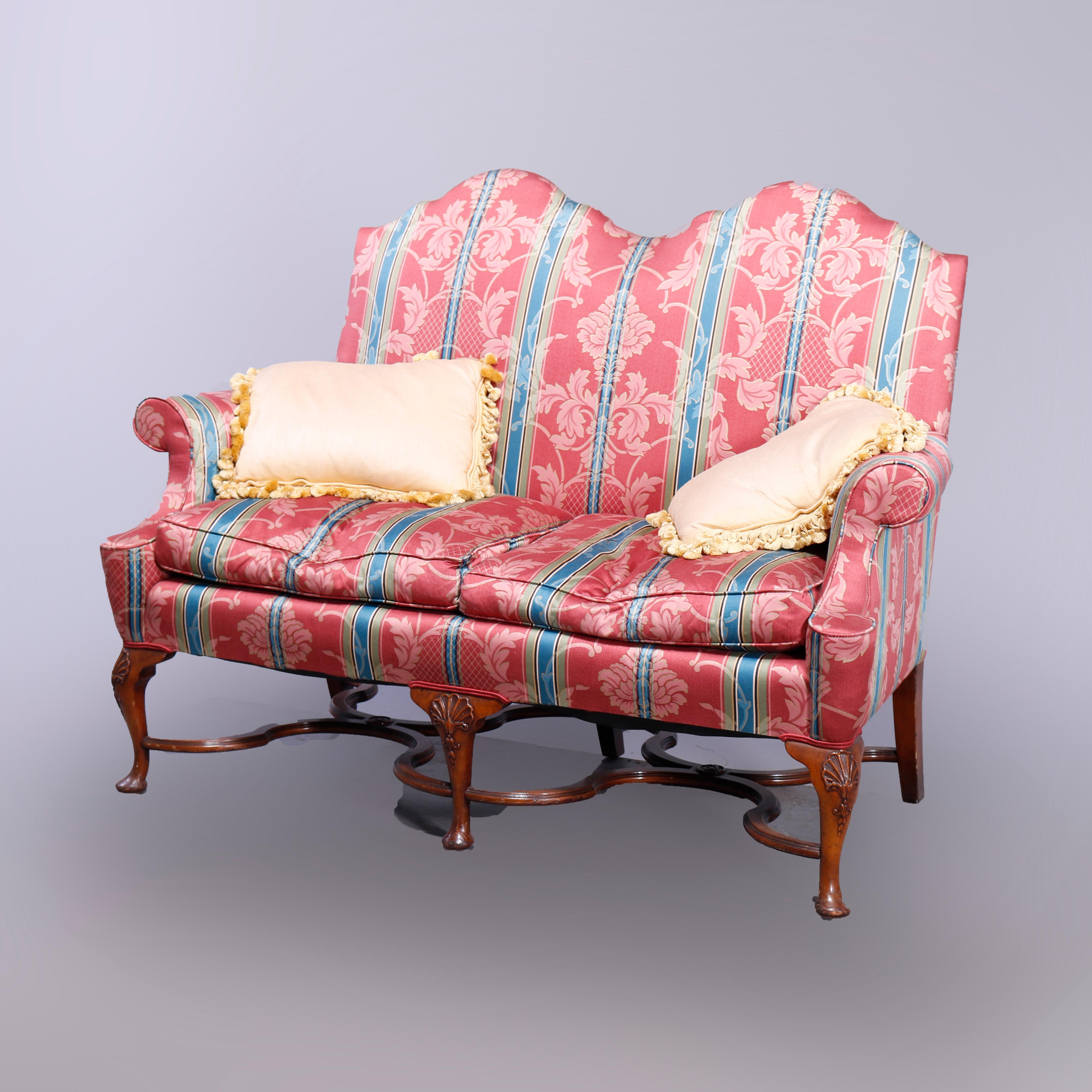 An English Queen Anne style settee sofa offers upholstered double camel back form with scroll arms raised on carved mahogany cabriole legs with shell knees, scroll stretcher and terminating in pad feet, 20th century

Measures - 43'' H x 56.5'' W x