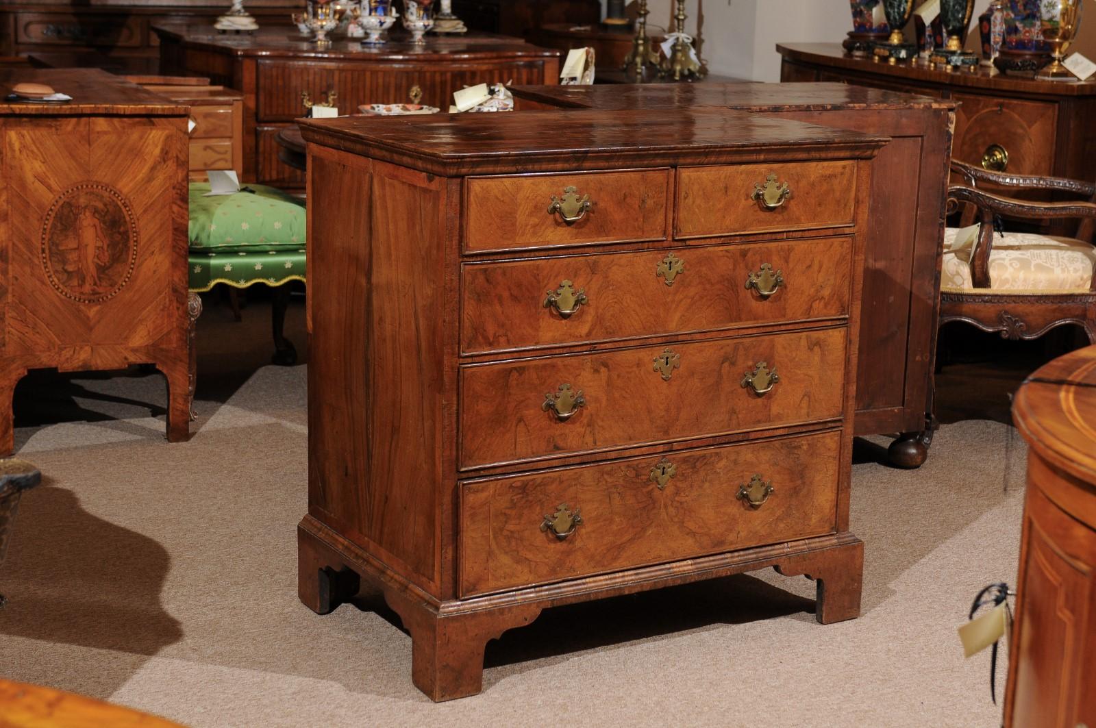 An early 18th century English Queen Anne period chest featuring figured walnut, cross-banding, 5 drawers with brass pulls ending on bracket feet.