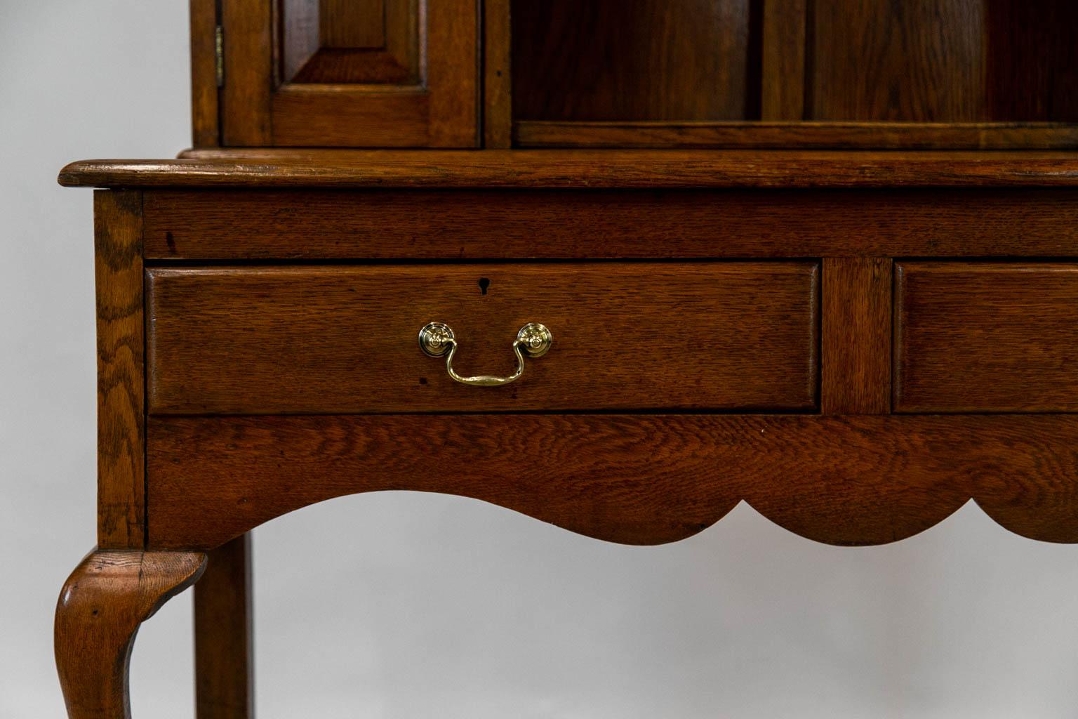 This oak welsh dresser has two doors in the top half that have raised gothic arch panels. The top has a scalloped frieze with three shelves that have a carved plate groove. The legs are cabriole and terminate in a slipper foot.