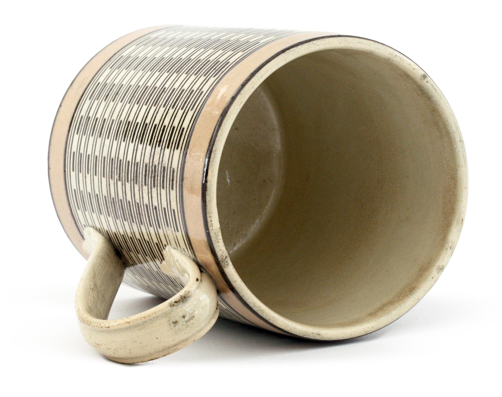 A rare and unusual antique mocha ware pottery mug the cream colored body decorated with black slip banding in a line and dash pattern set between two blush pink bands applied around the top and lower edges of the body. The mug has an ear shaped