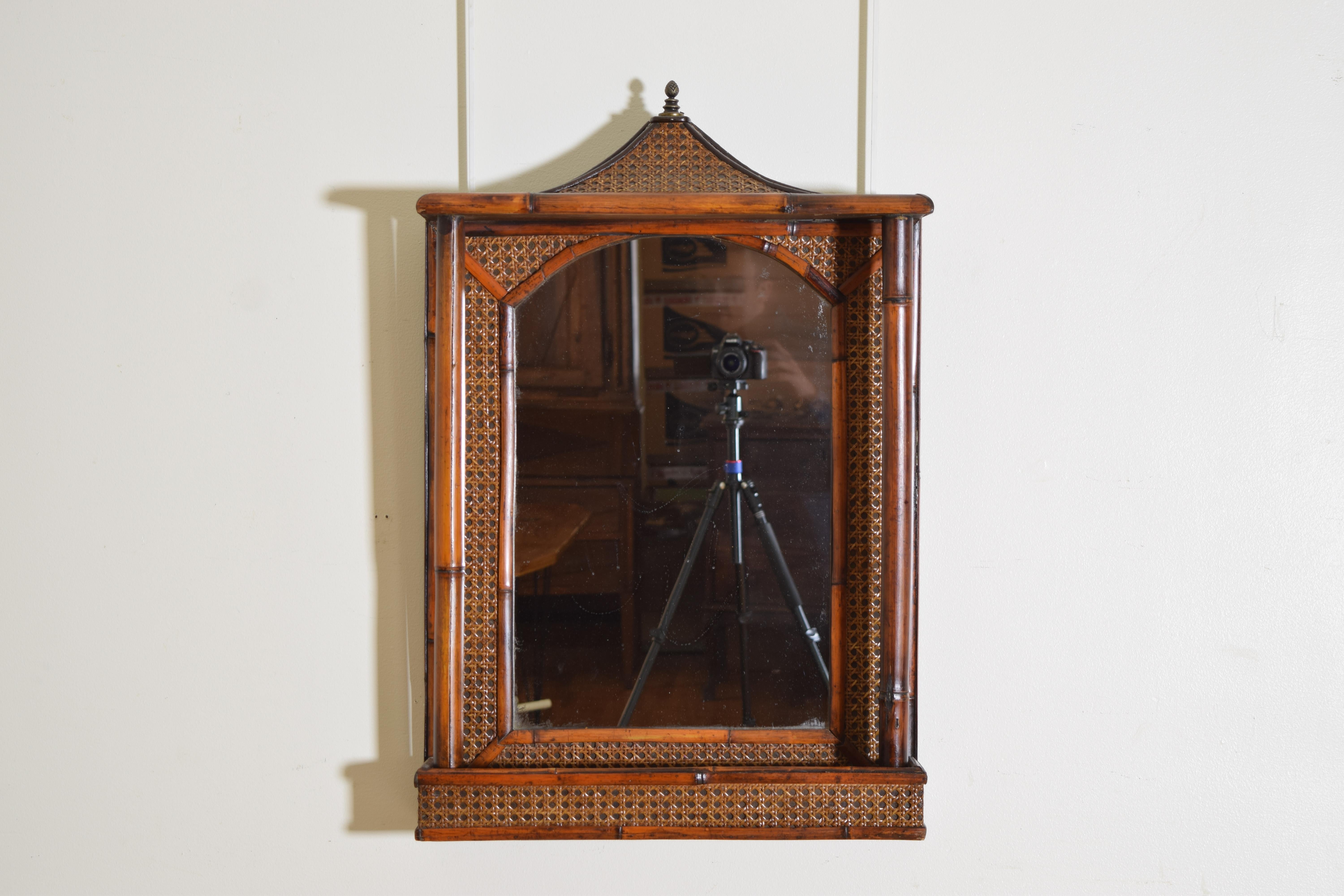 Having a pagoda shaped top with a cast brass finial and constructed of bamboo shaped wood with on a rattan ground, the mirror centered and behind a lower projected gallery used for storage in a bathroom