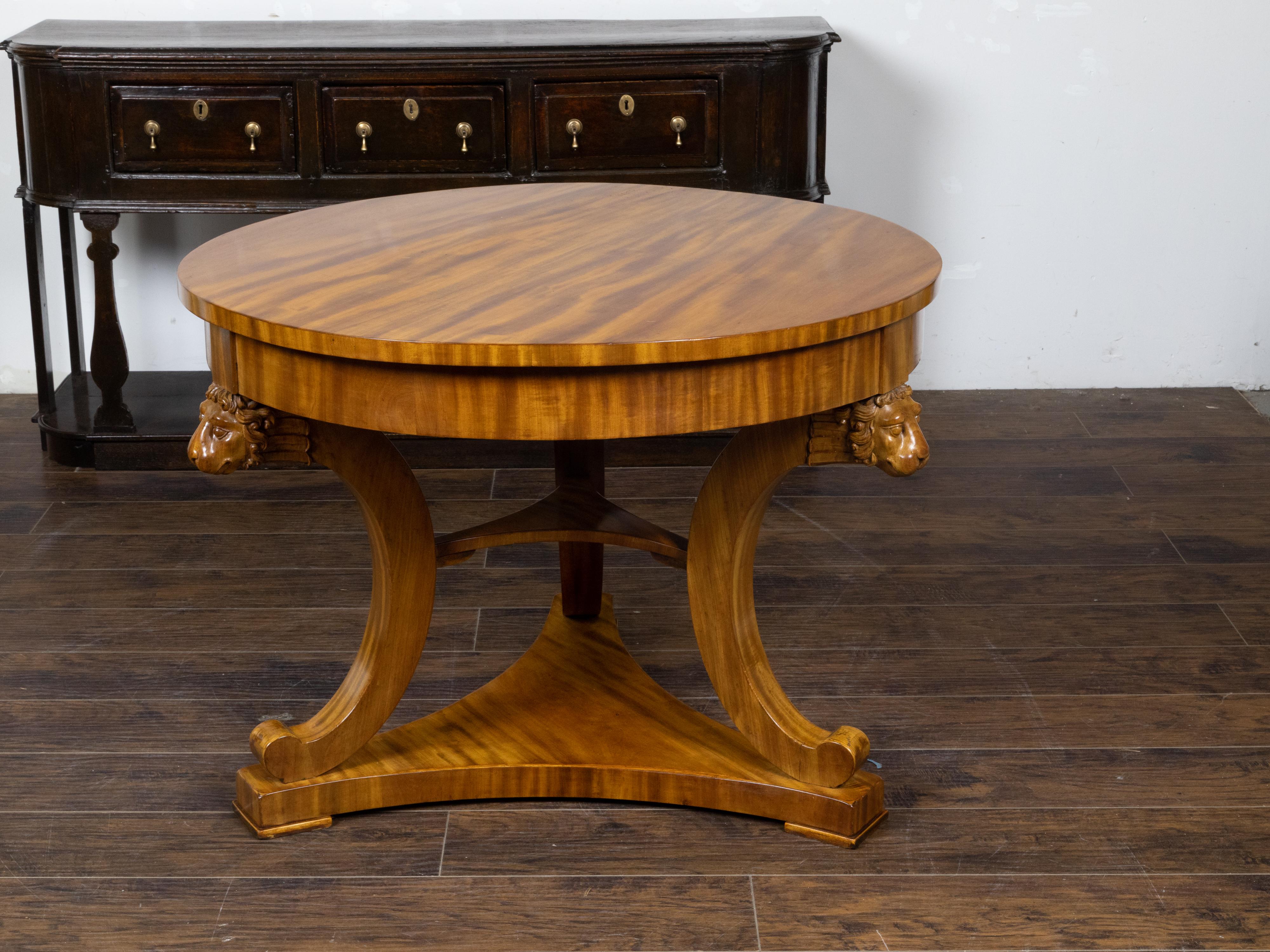 An English Regency period walnut center table from the 19th century, with round top, carved lion heads, tripod base and in-curving shelves. Created in England during the 19th century, this center table showcases the stylistic characteristics of the