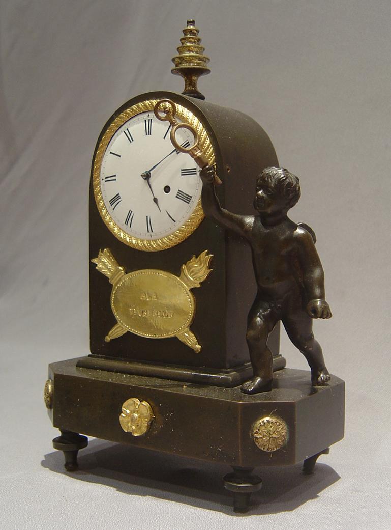 A small and unusual patinated bronze and ormolu English Regency mantel clock. Set on four feet the shaped base with ormolu mounts. A putti stands beside the clock housing with his arm raised. The clock is surmounted by a gilt bronze finial. The
