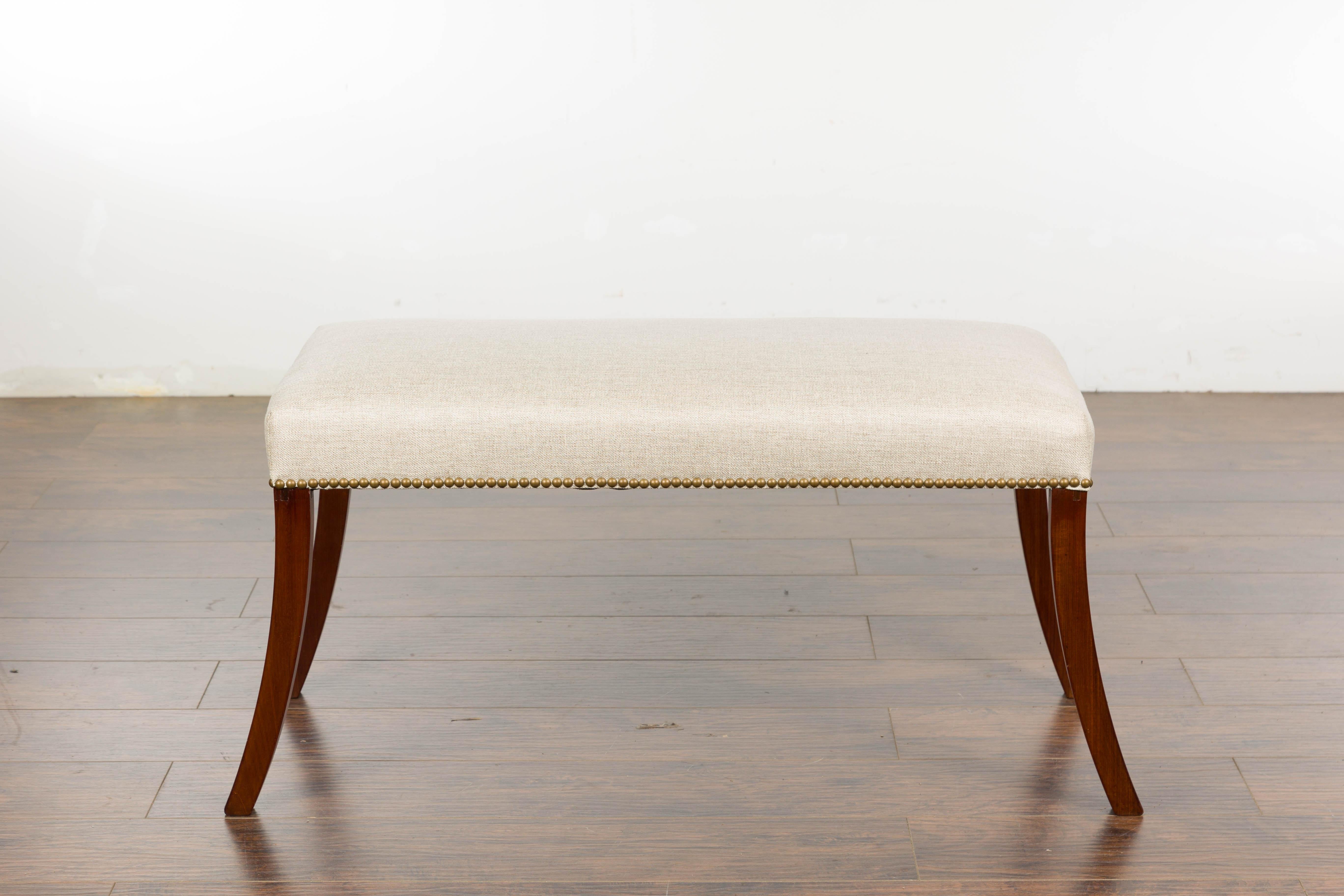 An English Regency bench from the 19th century with four saber legs and new custom linen upholstery with brass nailhead trim. Steeped in history and refined sophistication, this English Regency bench from the 19th century makes a stylish statement