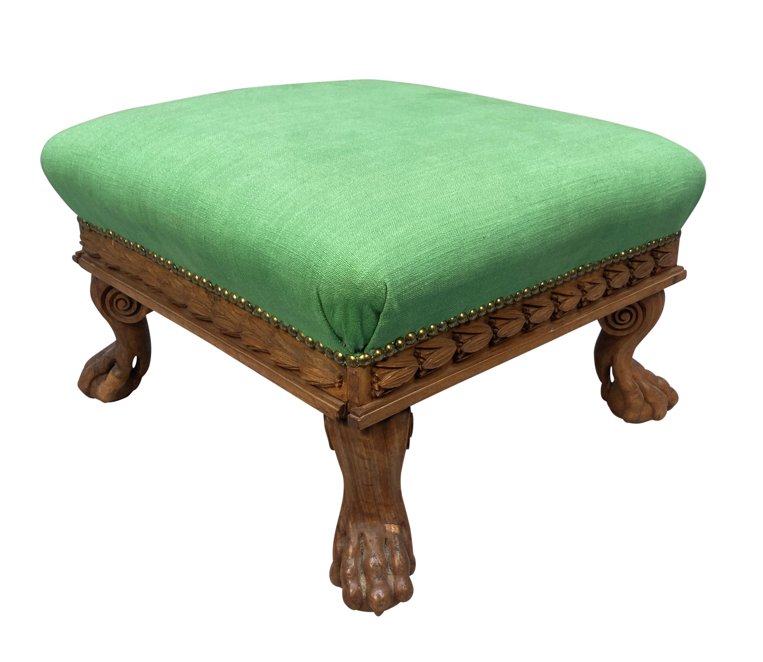 An English Regency, bleached mahogany footstool, crisply carved with a laurel frieze, and scrolled lion paw feet. Upholstered in a Fermoie linen.