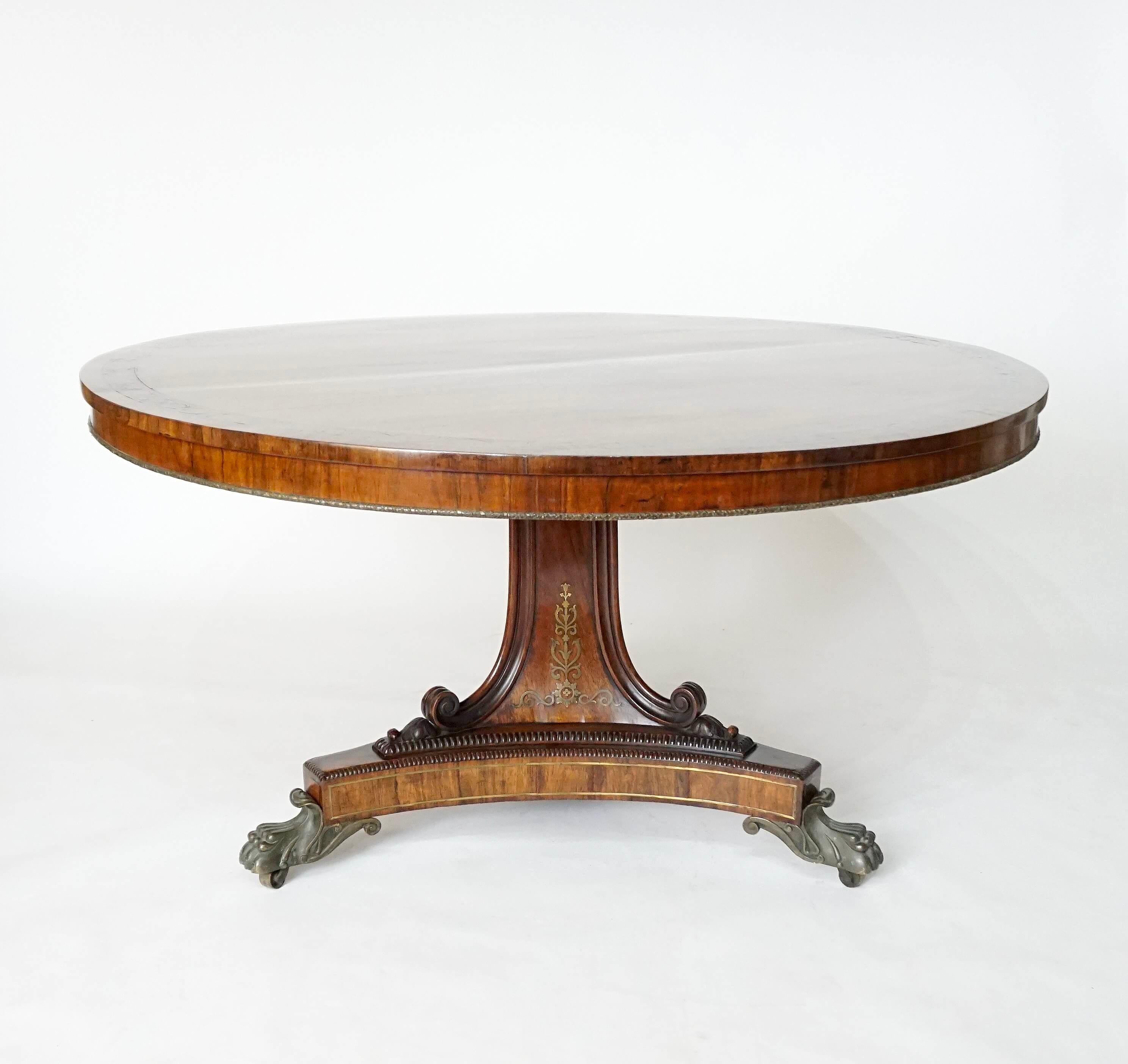 Elegant circa 1820 English Regency rosewood-veneered mahogany center table; the tilting top with foliate rinceau brass inlaid border with moulded brass ribbon-bound foliate garland bottom edge on foliate brass inlaid center trigonal pedestal support