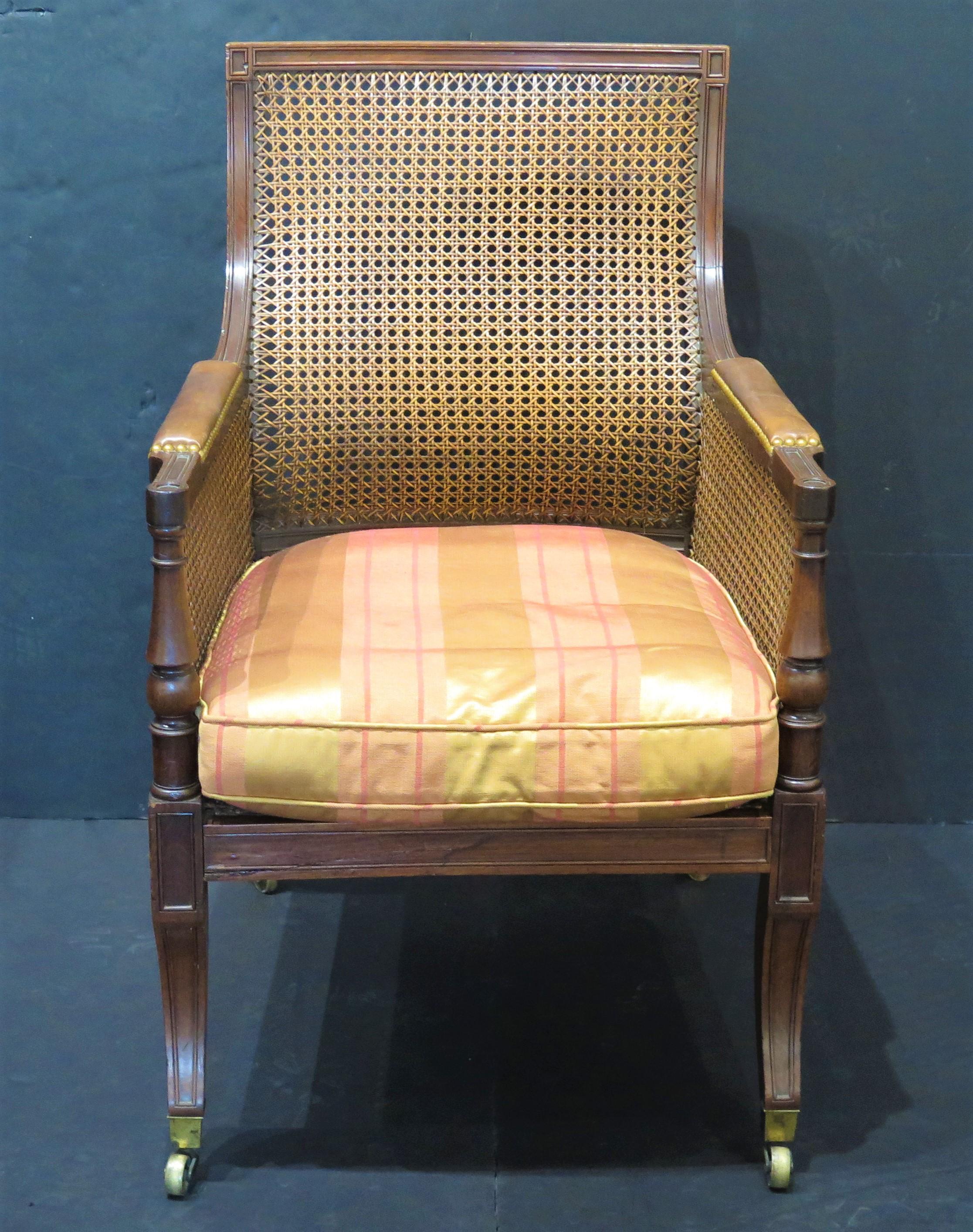 an English Regency caned library chair with loose cushion seat of striped silk, caned seat, back and sides, padded leather arm rests wi9th nailhead trim, sabre legs ending in brass caps with castors, England, mid-19th century