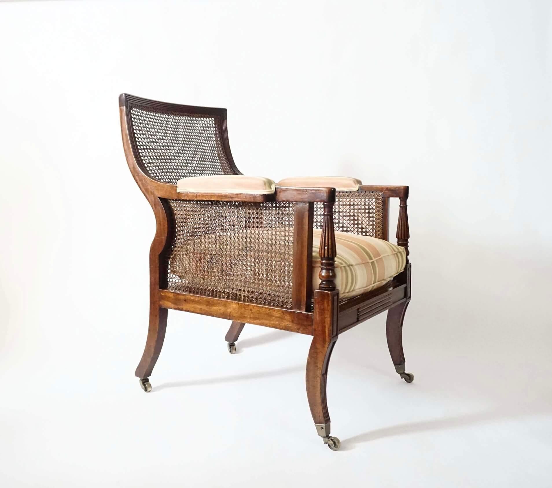 A fine and elegant English late George III library chair or bergère of large scale having solid mahogany frame with caned back, sides, and deep seat with loose squab cushion, the shaped and reclined back with reeded crestrail and sides continuing