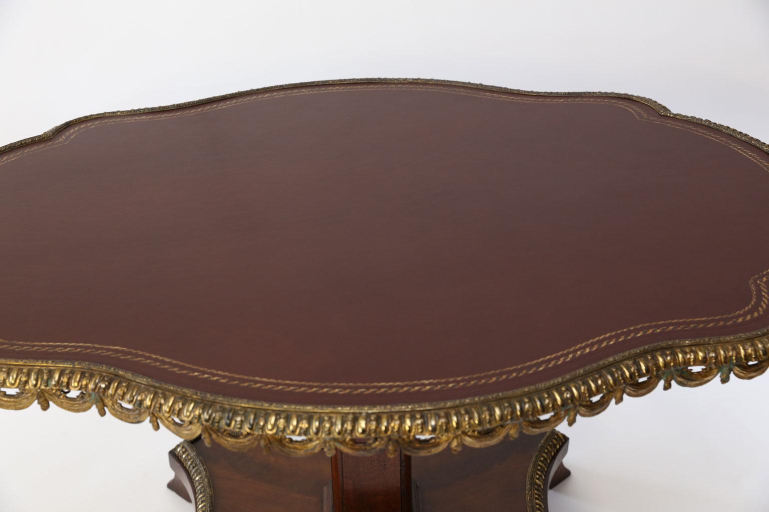 English leather-top center table in now-extinct Cuban mahogany. Leather-covered shaped top surrounded by original mercury-gilt cast brass decorative edge. Leather top is hand-tooled and gilded. Base features matching mercury-gilt cast brass