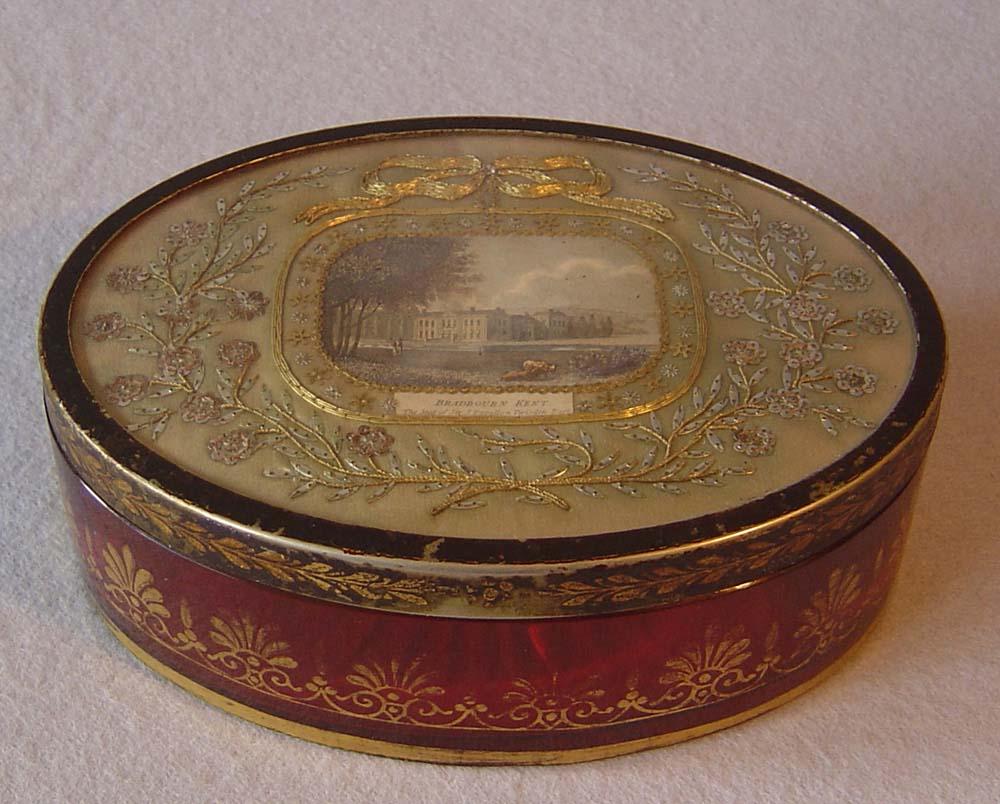 A magnificent English Regency oval cranberry glass dressing table box with toleware lid inset with embroidery surrounding a colour wash print. The cranberry glass with deep cut diamond shape to base and decorated around the sides in gilt amthemions.