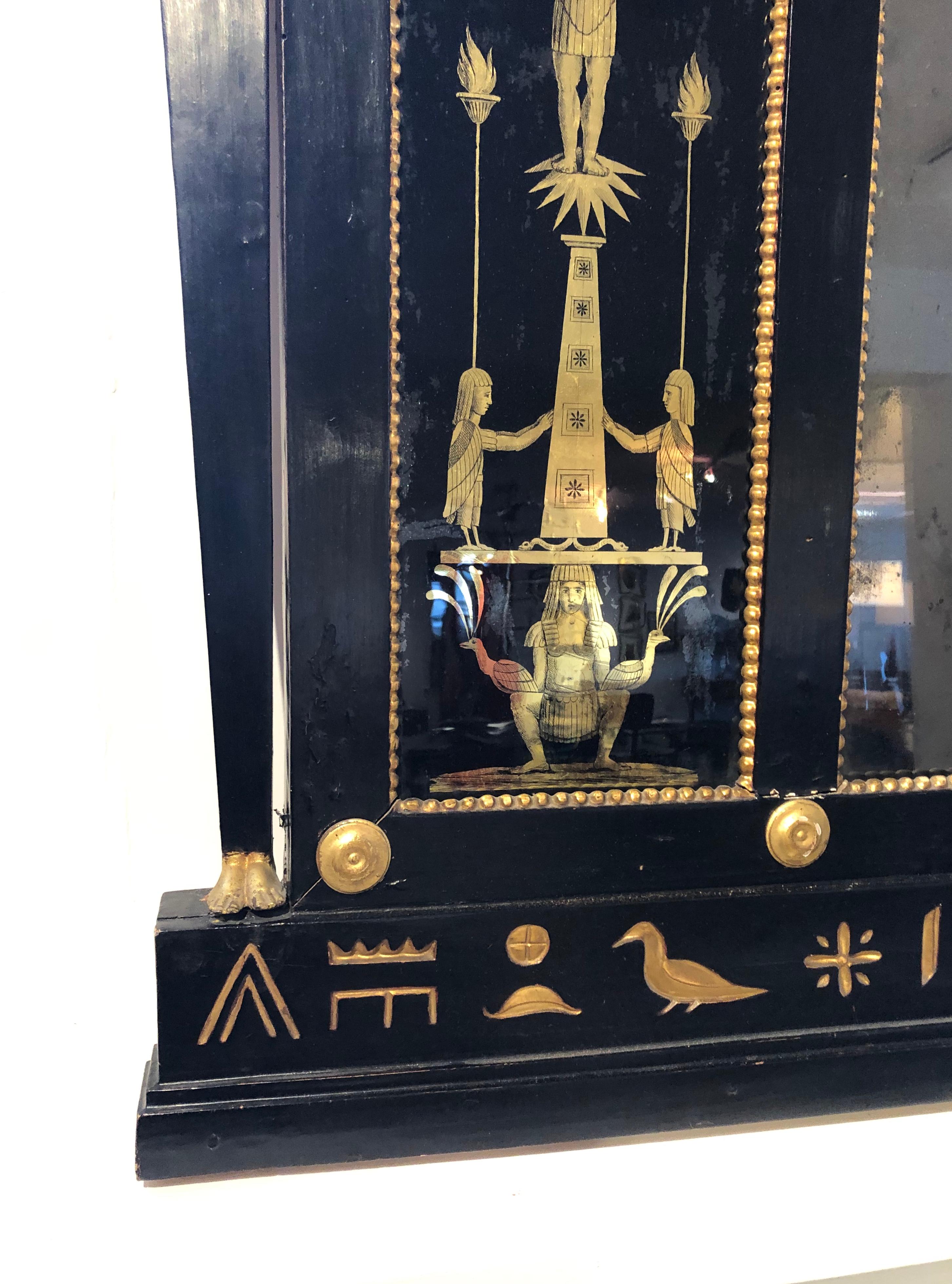 An English Regency neoclassical Egyptian Revival overmantel mirror, ebonized and parcel-gilt, of rectangular form, with pilasters on each side with gilded pharaoh's heads and feet, flanking black and gold reverse painted glass panels and with gilded