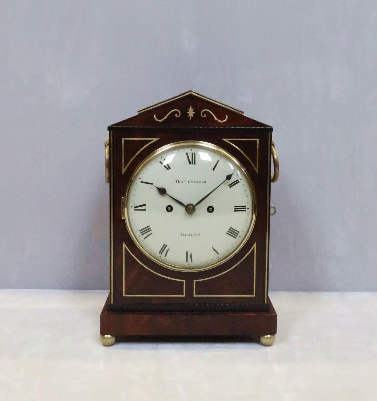 An extremely good quality English figured mahogany bracket or table clock with architectural shaped top and brass string inlay detail to the front, fish scale sound frets to the side and decorative ringed carrying handles stood on brass ball feet.
