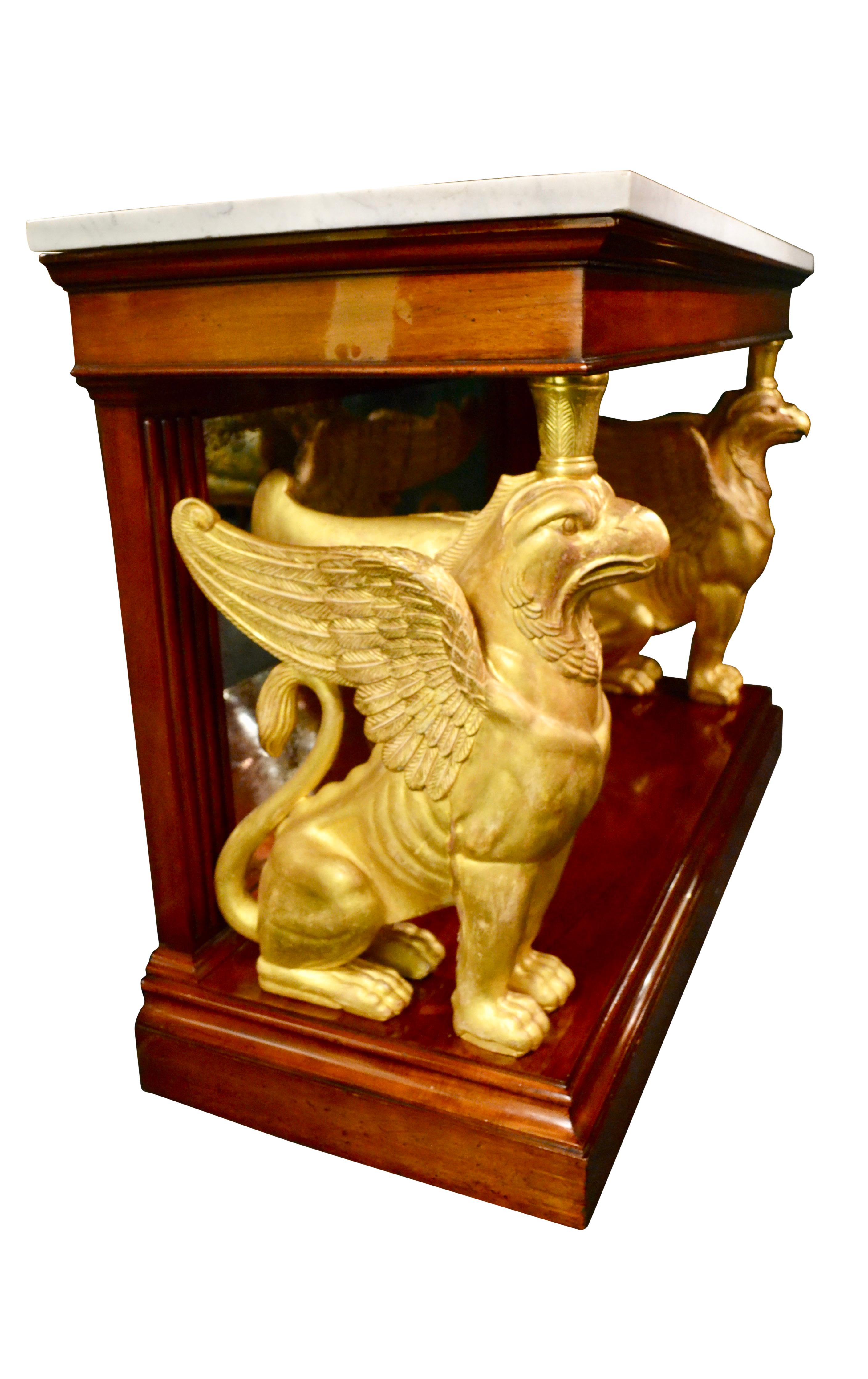 A large and impressive English Regency console table in mahogany and gilded wood; the top section with a thick white marble top rests on two large gilded winged wood gryphons ( griffins) joining to a lower mahogany base; the back of the console