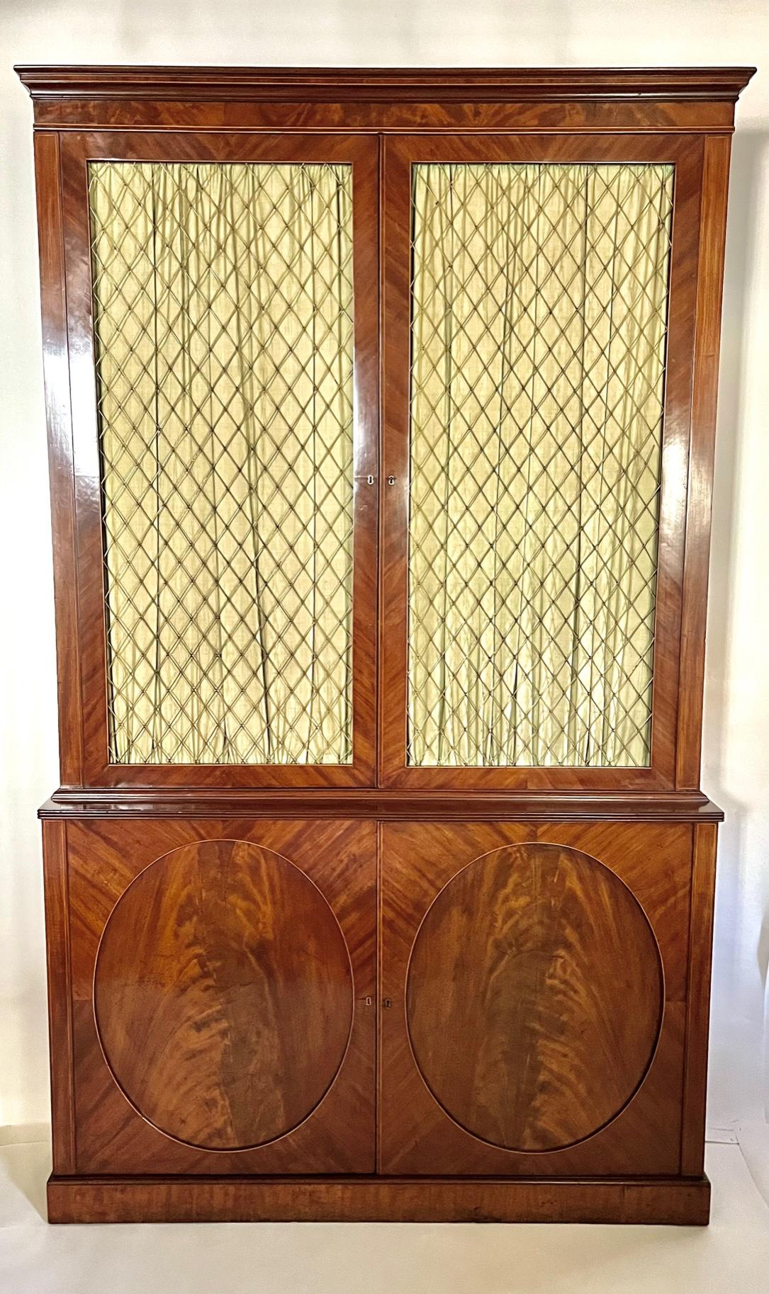 A fine and elegant circa 1810 English Late George III Regency period press cabinet of large scale having exquisitely grained mahogany veneered and satinwood inlaid case, the fixed moulded cornice surmounting double harlequin or lozenge pattern wire