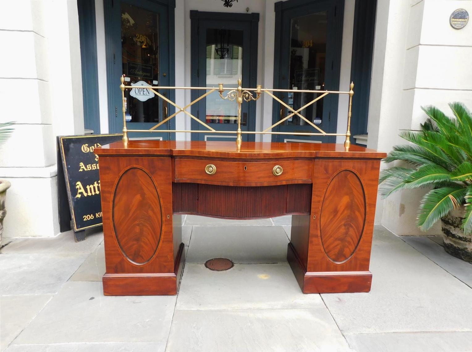 English Regency mahogany bow front sideboard with crosshatched bulbous urn finial gallery, centered scrolled acanthus floral candelabra, flanking oval book matched cabinet doors with fitted interior drawers, centered sliding tambour compartment,