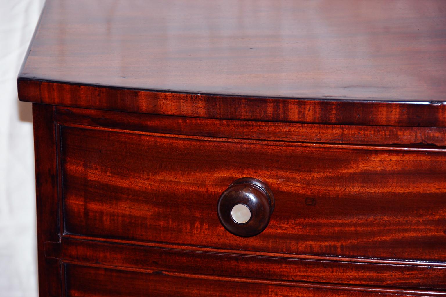 English Regency period mahogany bowfront chest of four graduated drawers. This handsome small chest retains its original mahogany knobs with mother of pearl inlaid centers, french feet and shaped skirt. The quality of the timber and craftsmanship