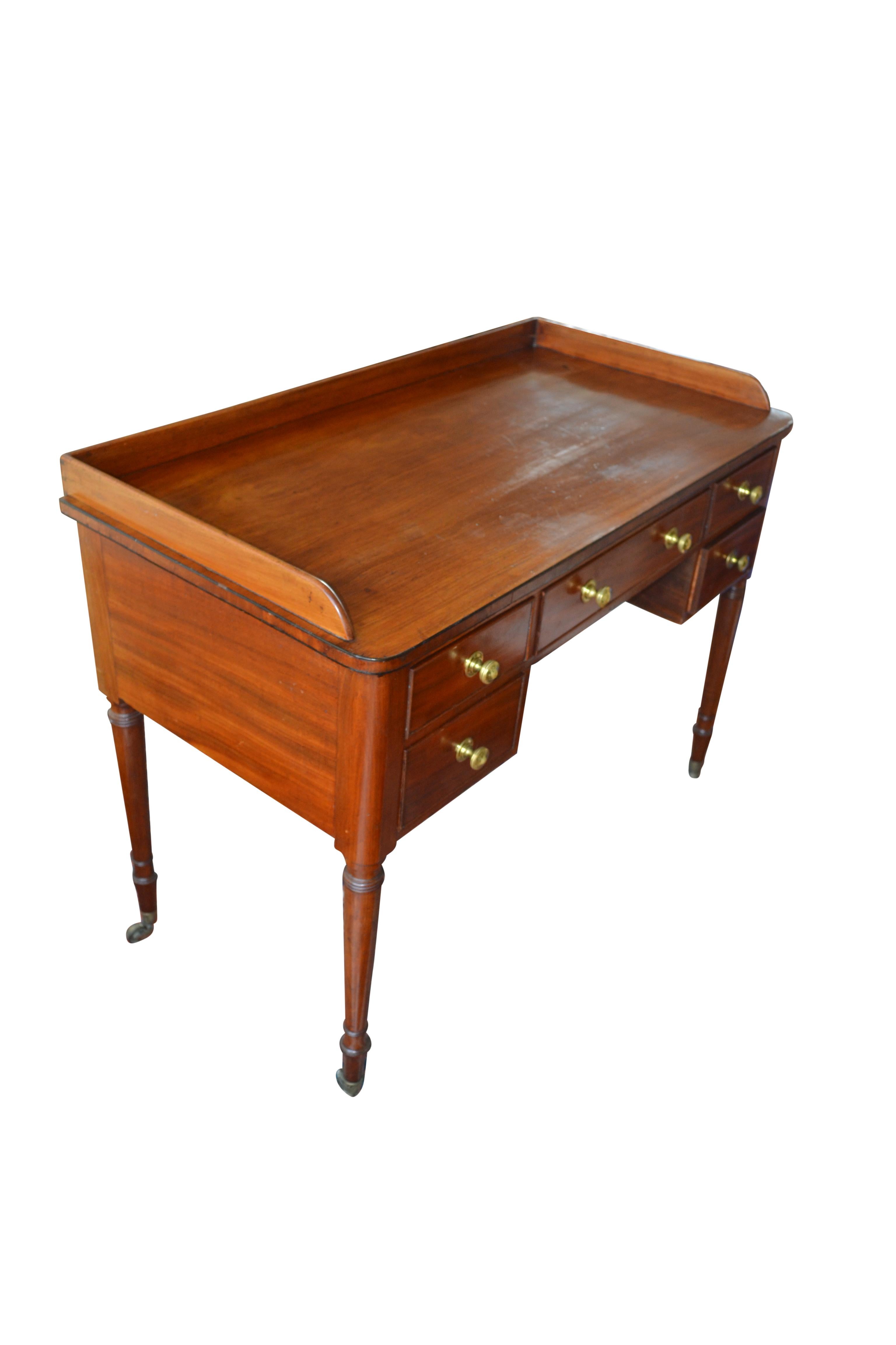 A finely figured English Regency mahogany desk/table/dressing table/sideboard with a three quarter wooden gallery over five drawers with their original brass hardware. The table rests on four turned and tapered legs also with their original brass