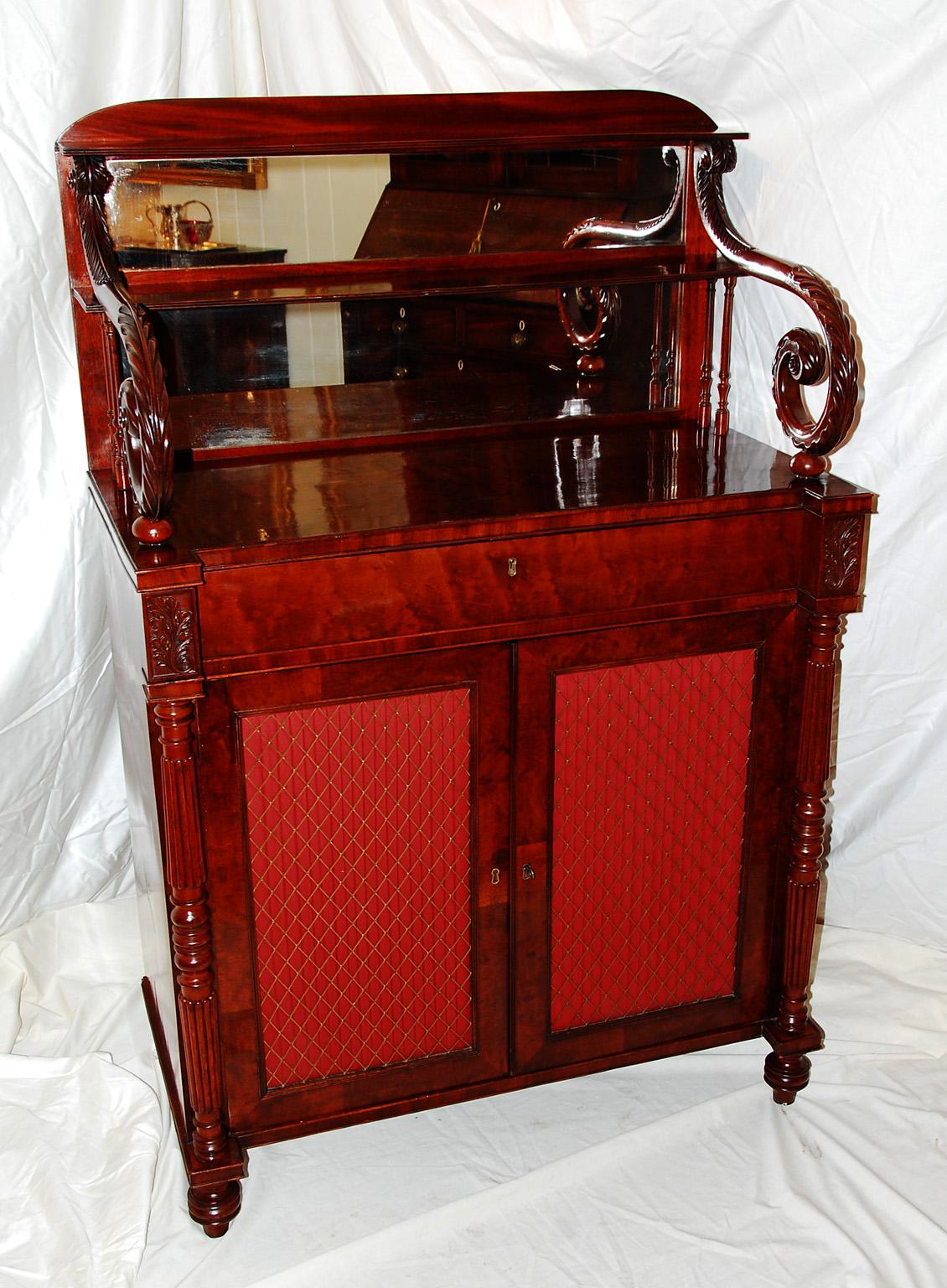 English Regency period flame grained mahogany chiffoniere with mirrored back, and two open shelves flanked by finely carved acanthus leaf scrolls. The two cabinet doors are inset with brass grilles, behind which is pleated fabric (which could easily