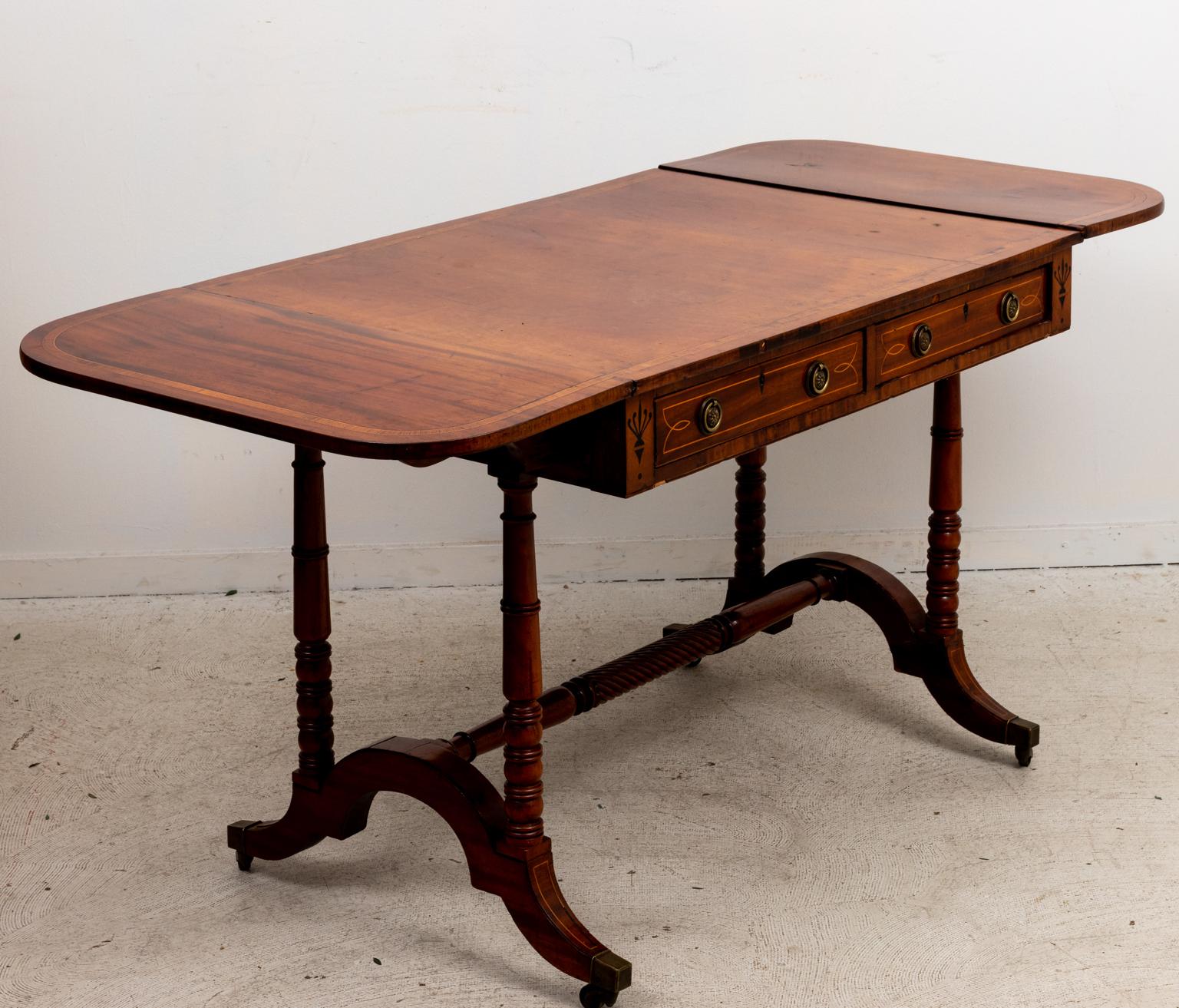Circa late 19th century English Regency style mahogany drop leaf sofa table on trestle base with castors. The desk also features inlaid trim on the drawer fronts and tabletop, ball-and-ring turned legs, and a central barley twist shaped bottom