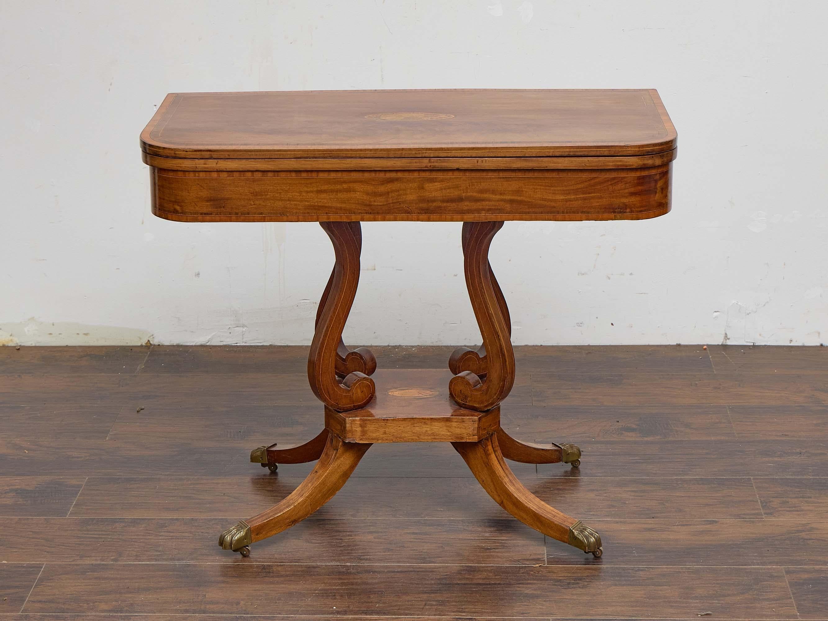 An English Regency mahogany games table, dating to the early 19th century, combining elegant form with practical versatility. The rectangular top, inlaid with a central satinwood medallion and crossbanding, swivels and opens to reveal a square