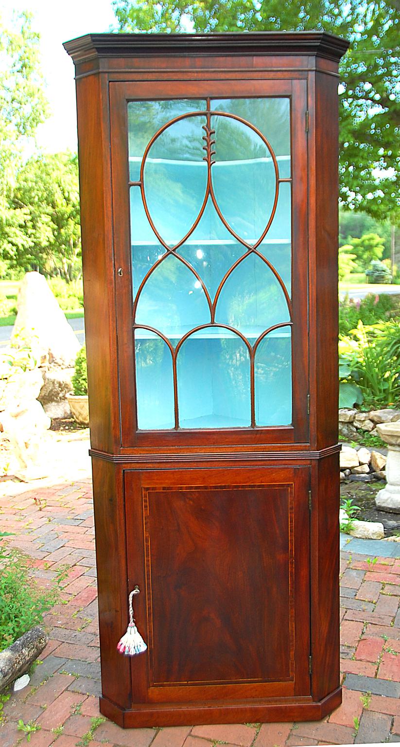 English Regency mahogany corner cupboard in two parts with the top having an astragal glazed and carved door in an unusual pattern, and an ogee cornice. The bottom has a paneled door with mahogany crossbanding enclosed by boxwood stringing, on a