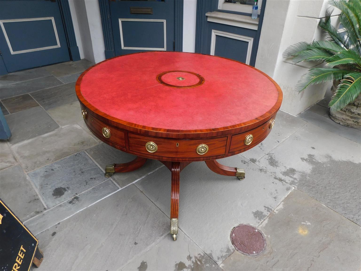 English Regency mahogany circular rotating leather top rent table with centered removable deposit lid, alternating four drawers, brass pulls, ebony string inlay, supported by a turned ringed pedestal resting on four reeded saber legs with the
