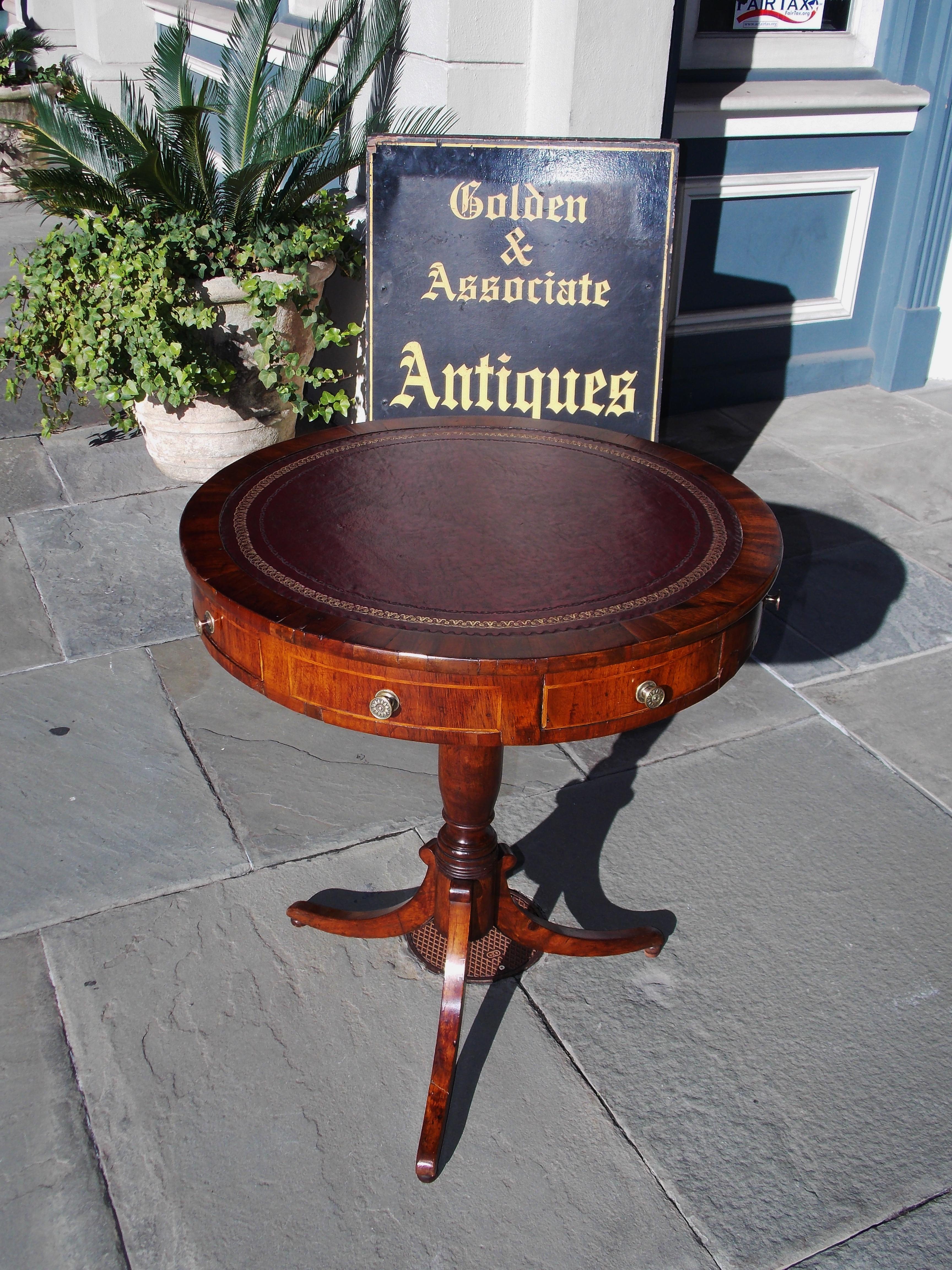 English Regency mahogany circular drum table with a red leather top and gilt tooling, alternating satinwood inlaid drawers with period floral brasses, carved turned bulbous ringed pedestal , and terminating on the original splayed legs with ball