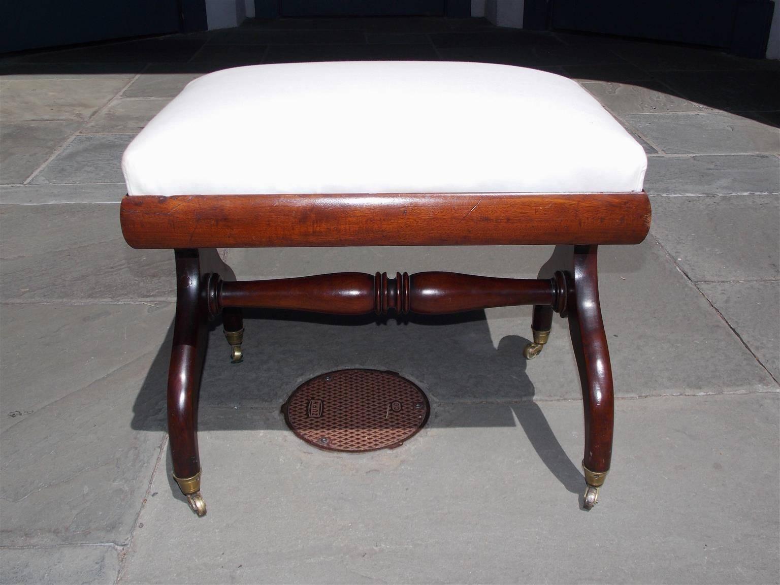 English Regency mahogany stool with a carved molded edge, stylized floral motif, turned bulbous ringed stretcher, and terminating on scrolled legs with the original brass casters. Stool is upholstered in white muslin, Early 19th century.