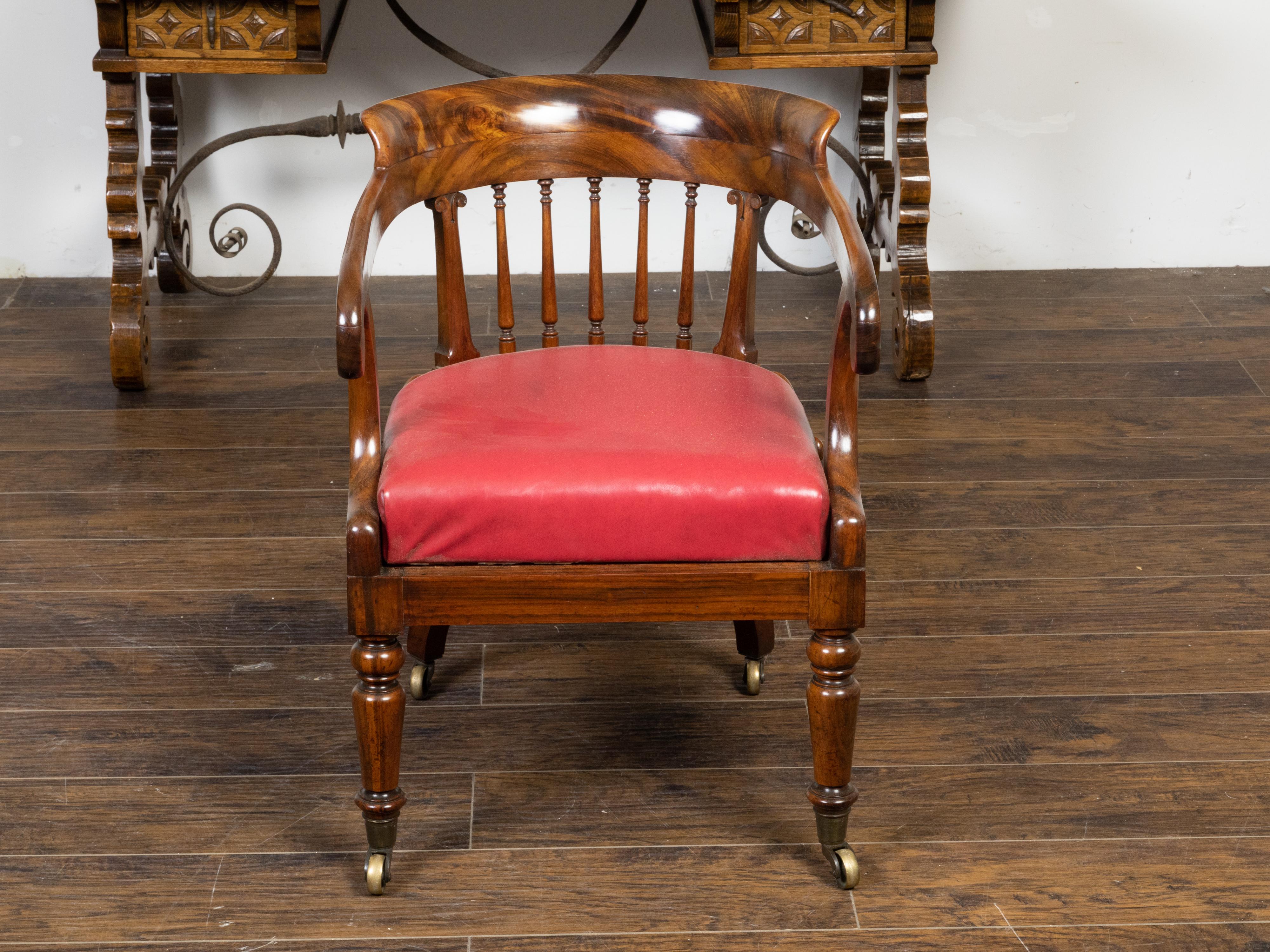 An English Regency period mahogany horseshoe back armchair from the 19th century, with balusters, large scrolling arms, carved volutes, turned legs, petite casters and red upholstery. Created in England during the Regency period in the early years