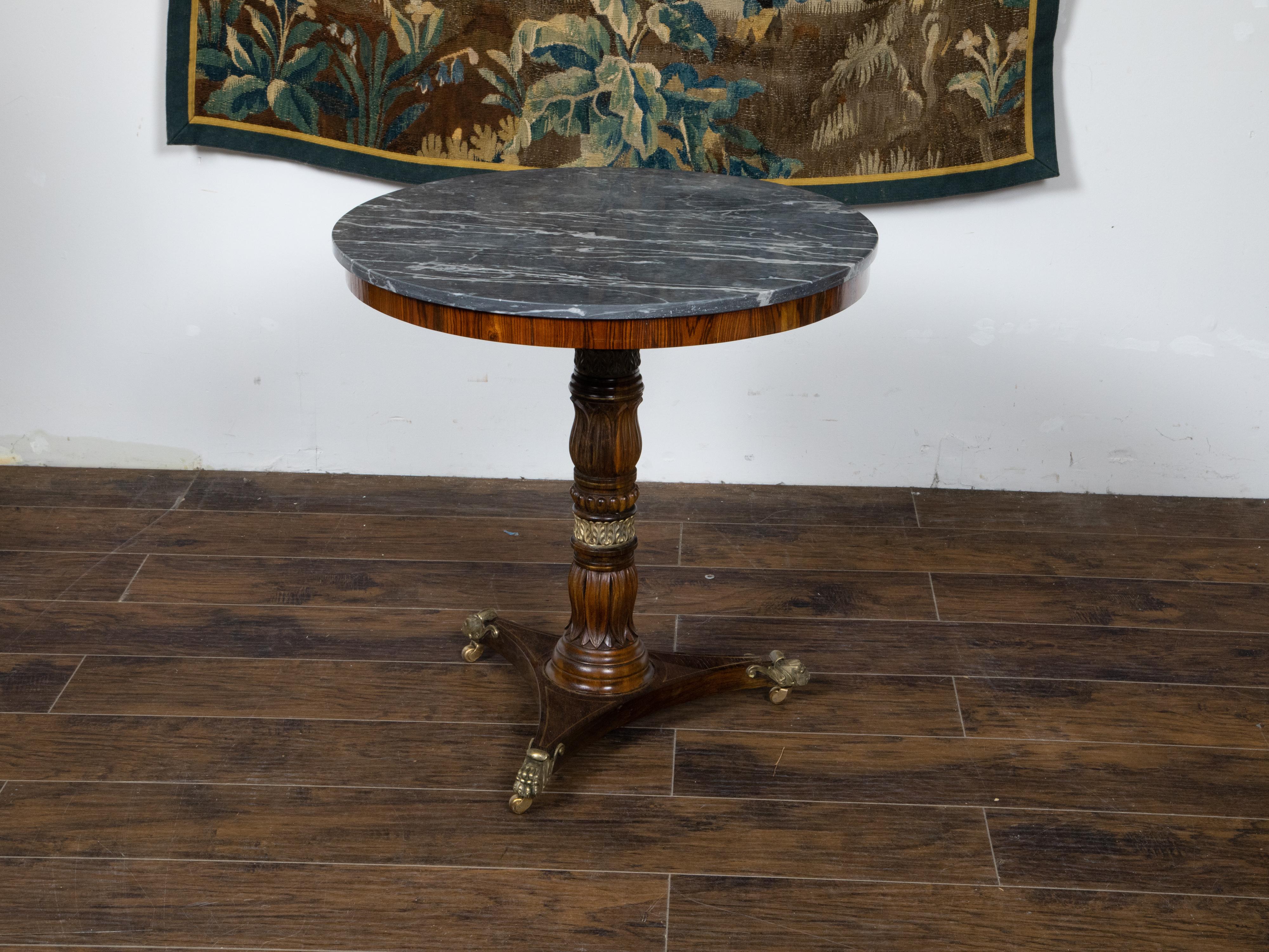 An English Regency period mahogany side table from the 19th century, with grey veined marble top, carved pedestal, tripod base and brass lion paw feet. Created in England during the Regency period, this mahogany side table features a circular top
