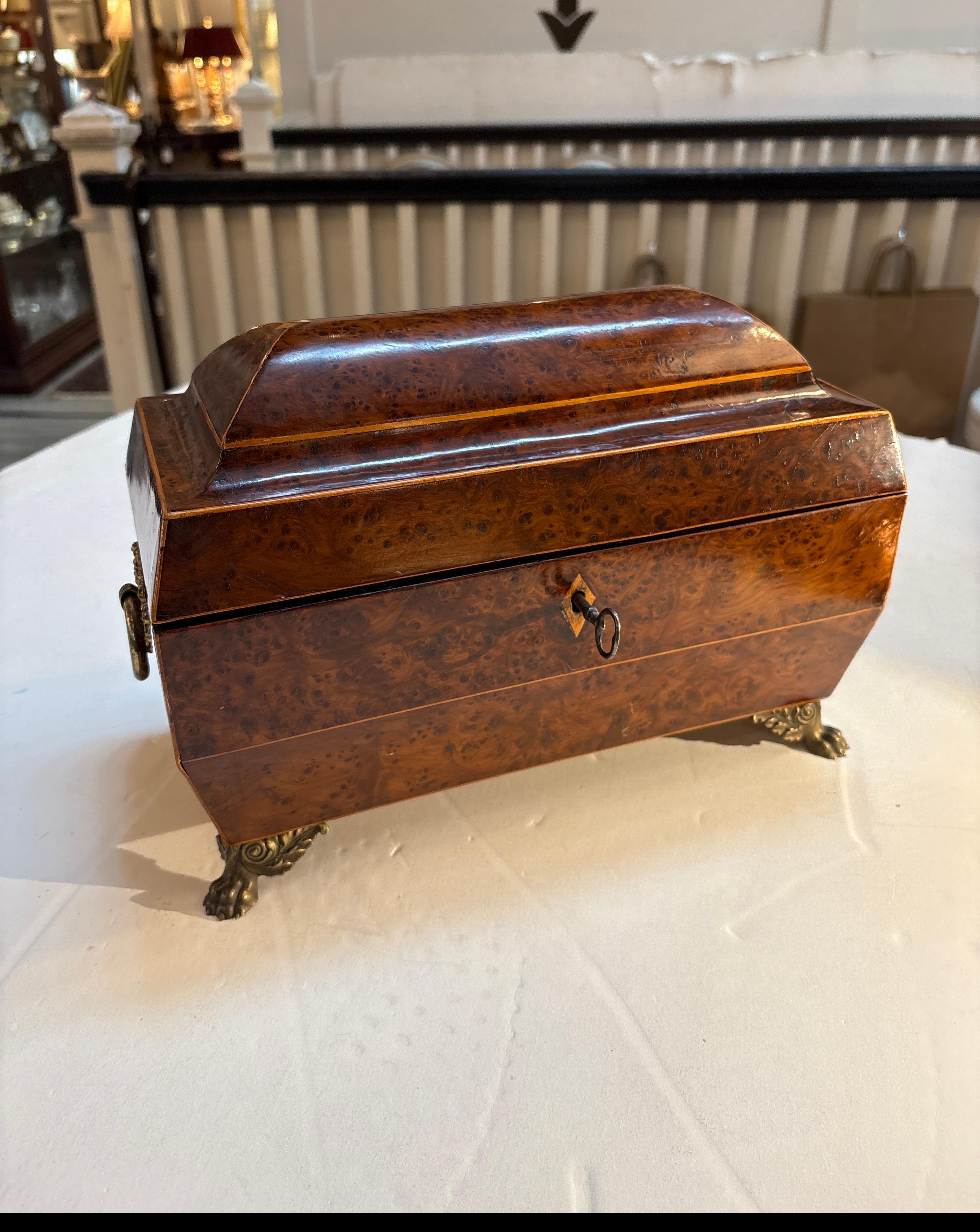 Un plateau à thé en amboyna marqueté de style Régence anglaise, avec des montures en laiton.  La forme de sarcophage avec des poignées de lion et des pieds à volutes.  L'intérieur comporte deux compartiments pour le thé avec un bol à mélanger en
