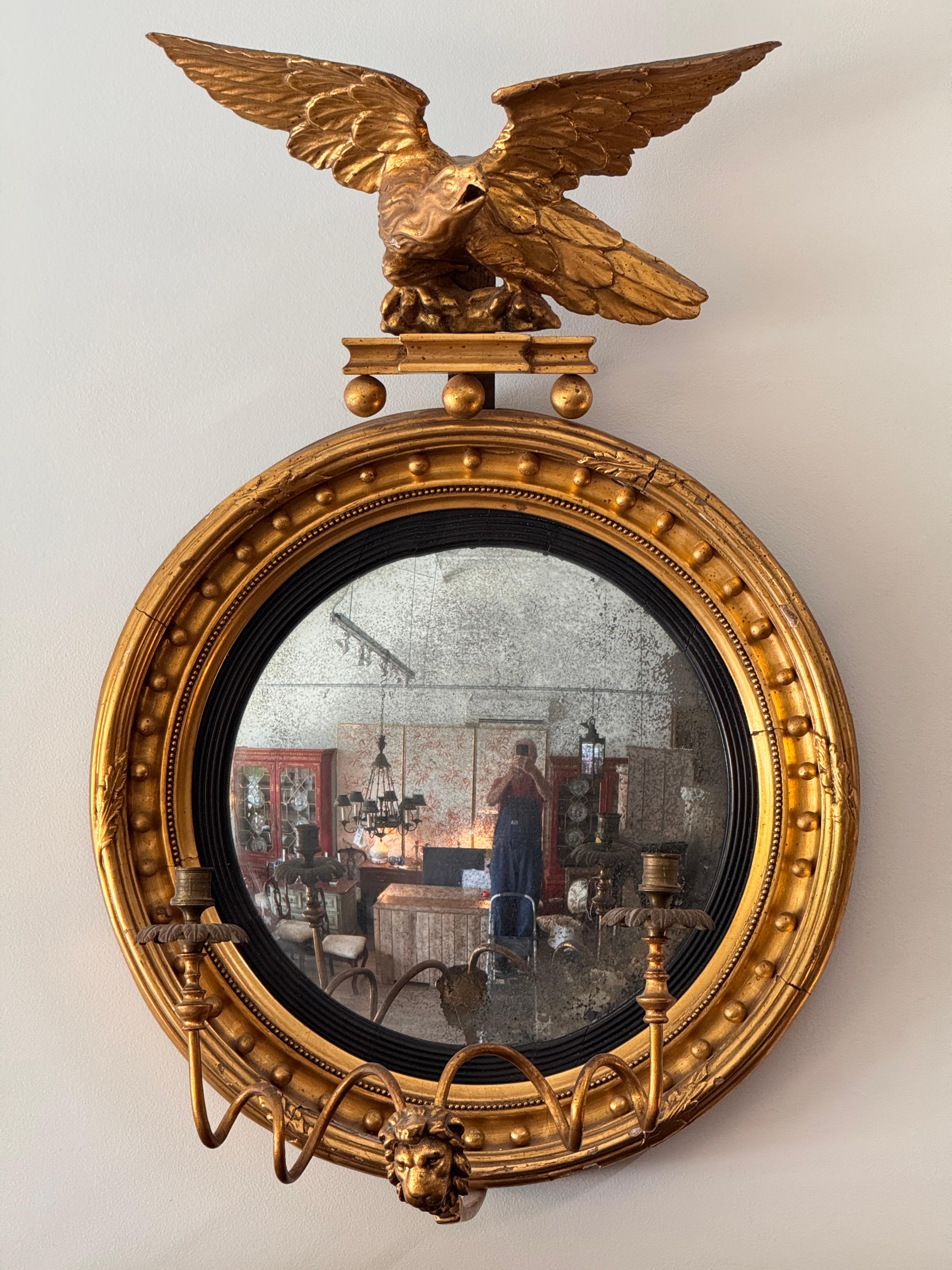 An English Regency period (c. 1810) giltwood and ebonized girandole convex mirror,  featuring candle arms and surmounted by a finely carved eagle. Beaded frame and lion mask base complete the classic design.
