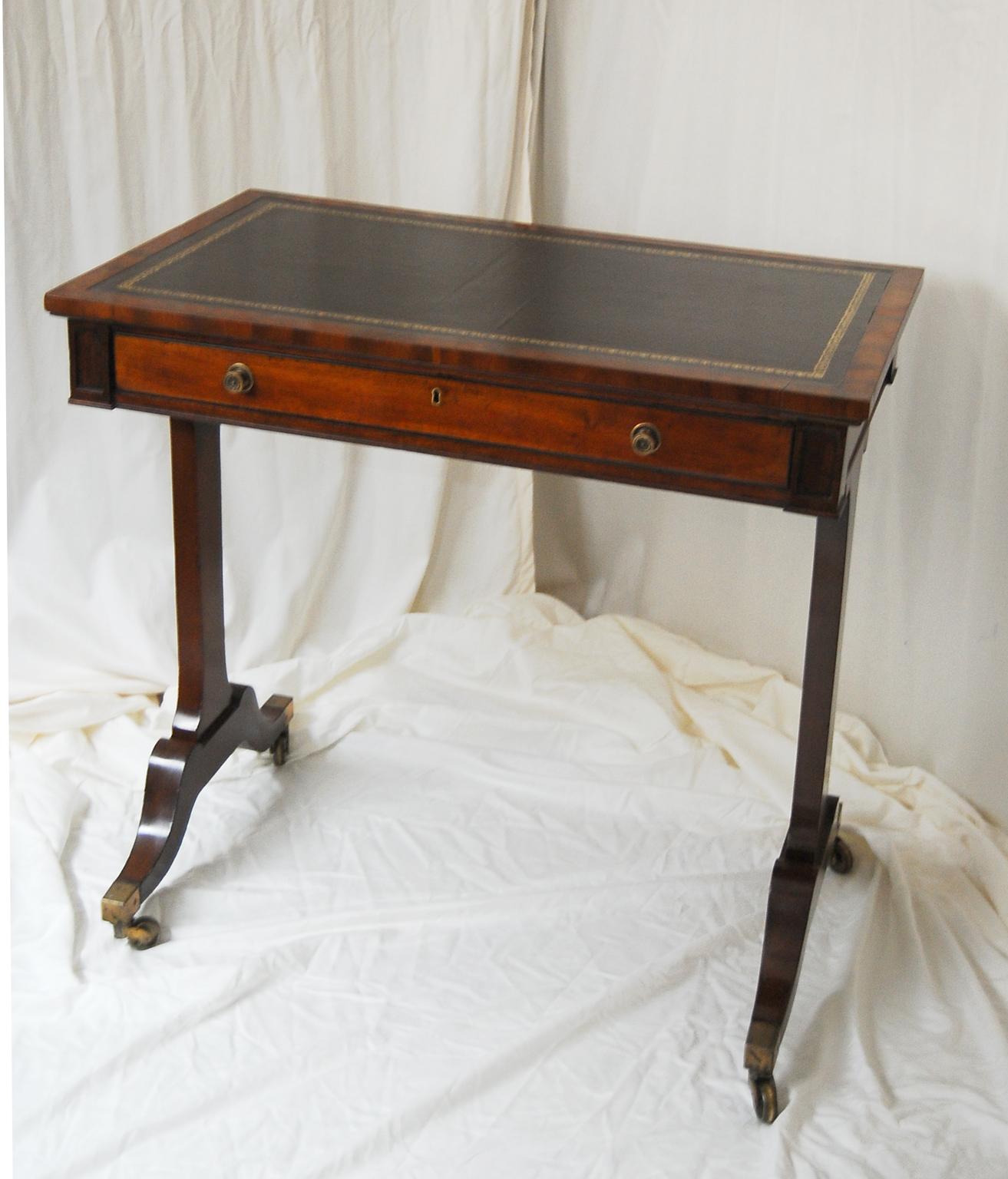English Regency period mahogany one drawer pedestal end writing table with black hand dyed and tooled leather writing surface. This small elegant writing table has downswept legs ending in brass toes and casters. It is finished on all four sides so