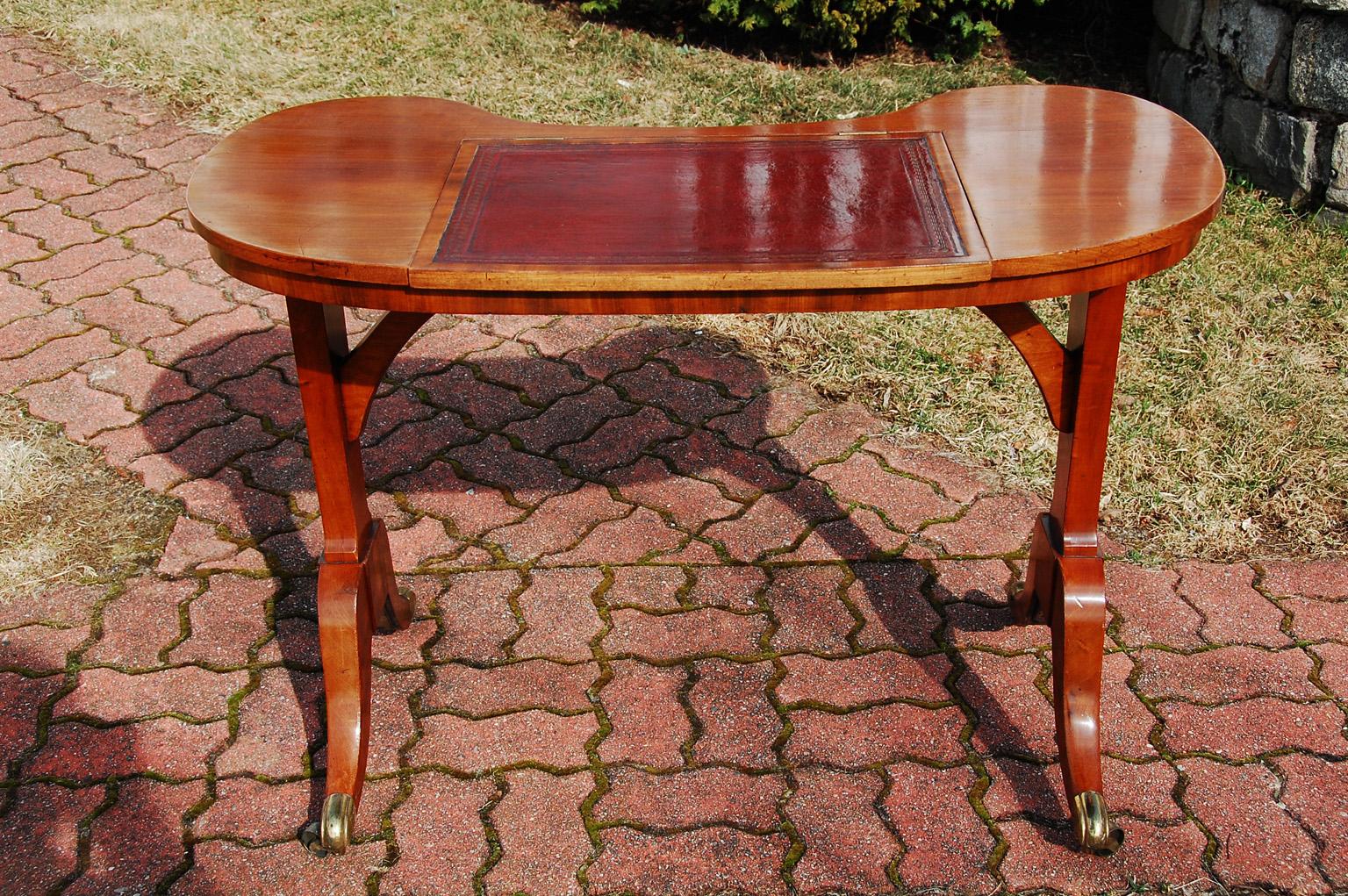 English Regency period satinwood kidney shaped writing table with pedestal ends and saber legs ending in brass casters. This elegant writing table has a hinged writing surface with inlaid hand tooled and hand dyed older red leather in excellent