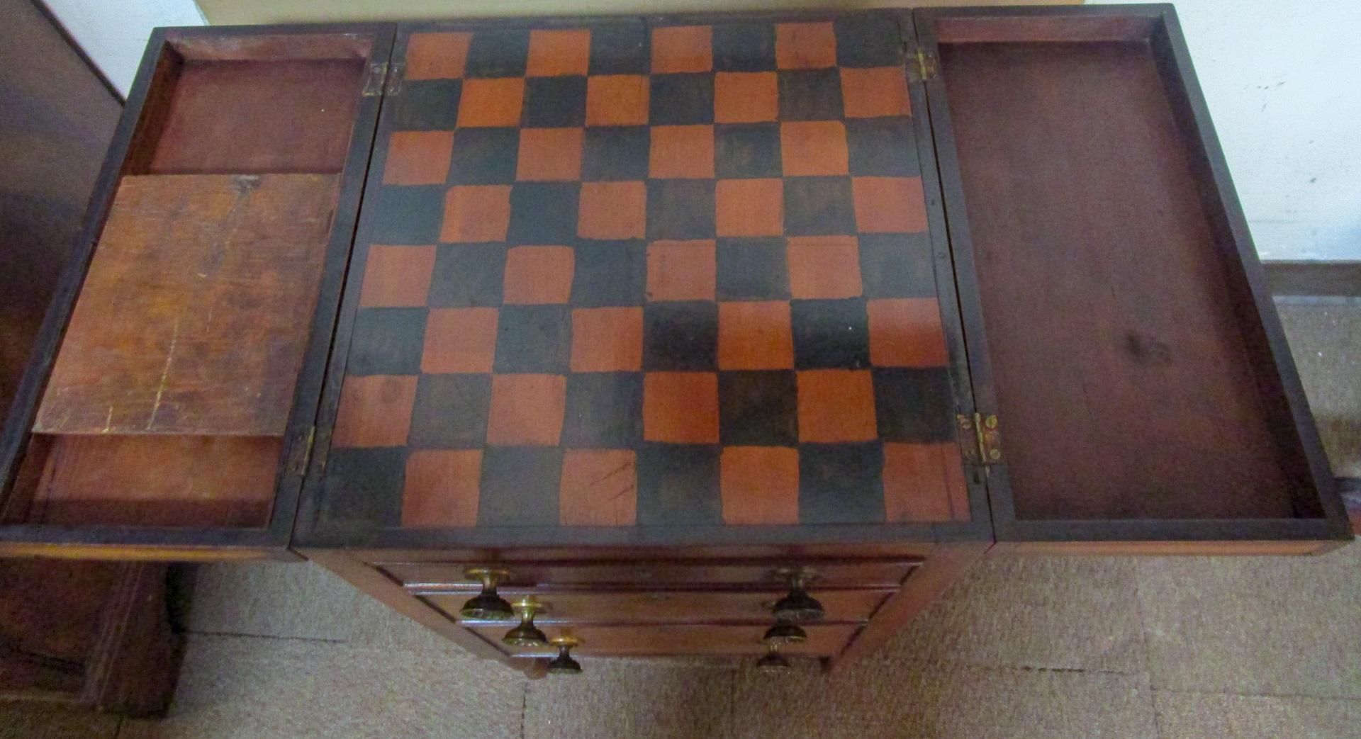 This unique petite size game table features three graduated sized drawers retaining the original brass fleurette motif pulls and a double hinged top that opens to a checkerboard. One of the folding sides contains a slider that stores game pieces and