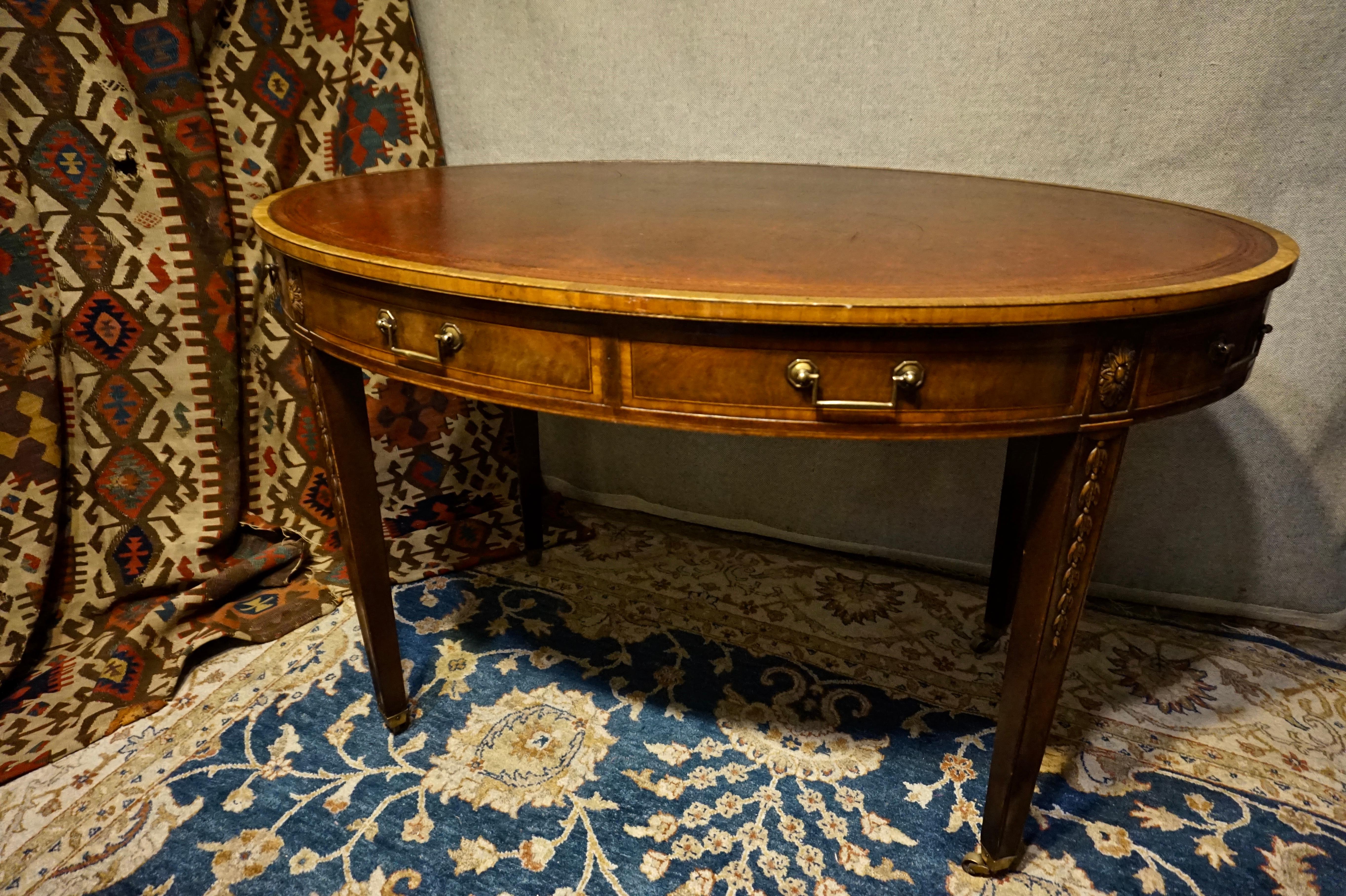 English Regency Revival oval leather writing table with old gilt laced leather top and brass hardware. Two drawers on one side with several mock drawers that wrap around the table. Hand carved. Casters at base. Solid wood. Excellent aesthetics. May